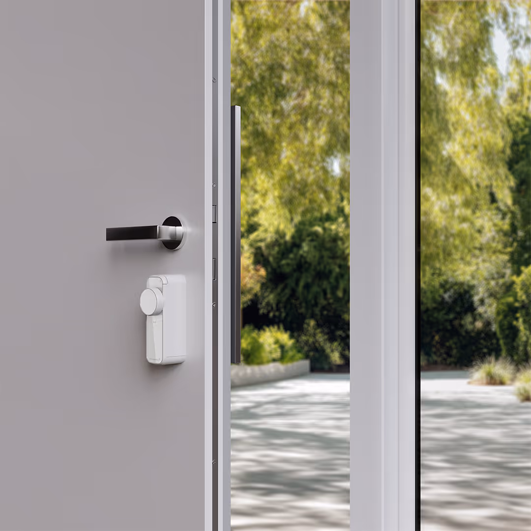 A modern gray door with a silver handle and a white smart lock, partially open showing a view of green trees outside.