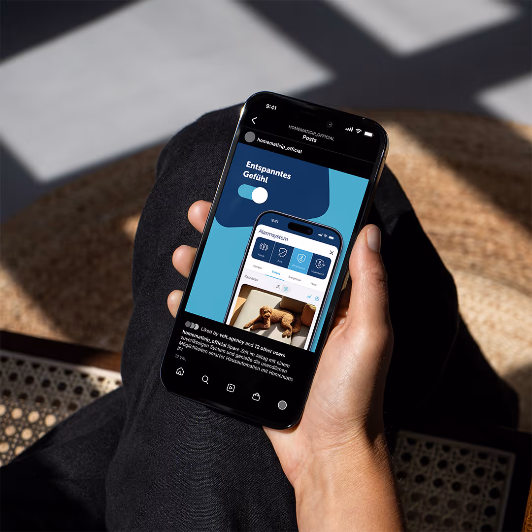 Hand holding a smartphone displaying a social media post about a home alarm system app showing a dog on a couch and German text.