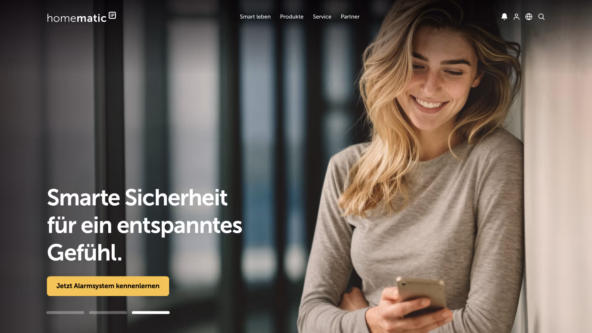 Smiling woman in gray shirt looking at her smartphone, leaning against a wall in a softly lit modern interior.