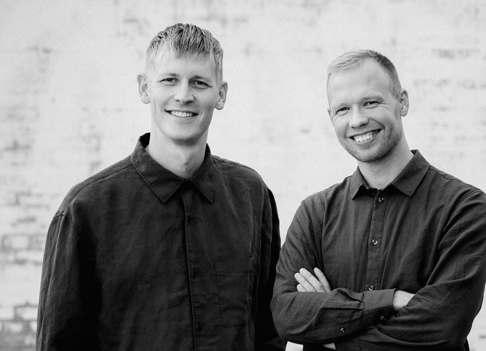 Two smiling men in dark shirts posing in front of a light brick wall, one with arms crossed.