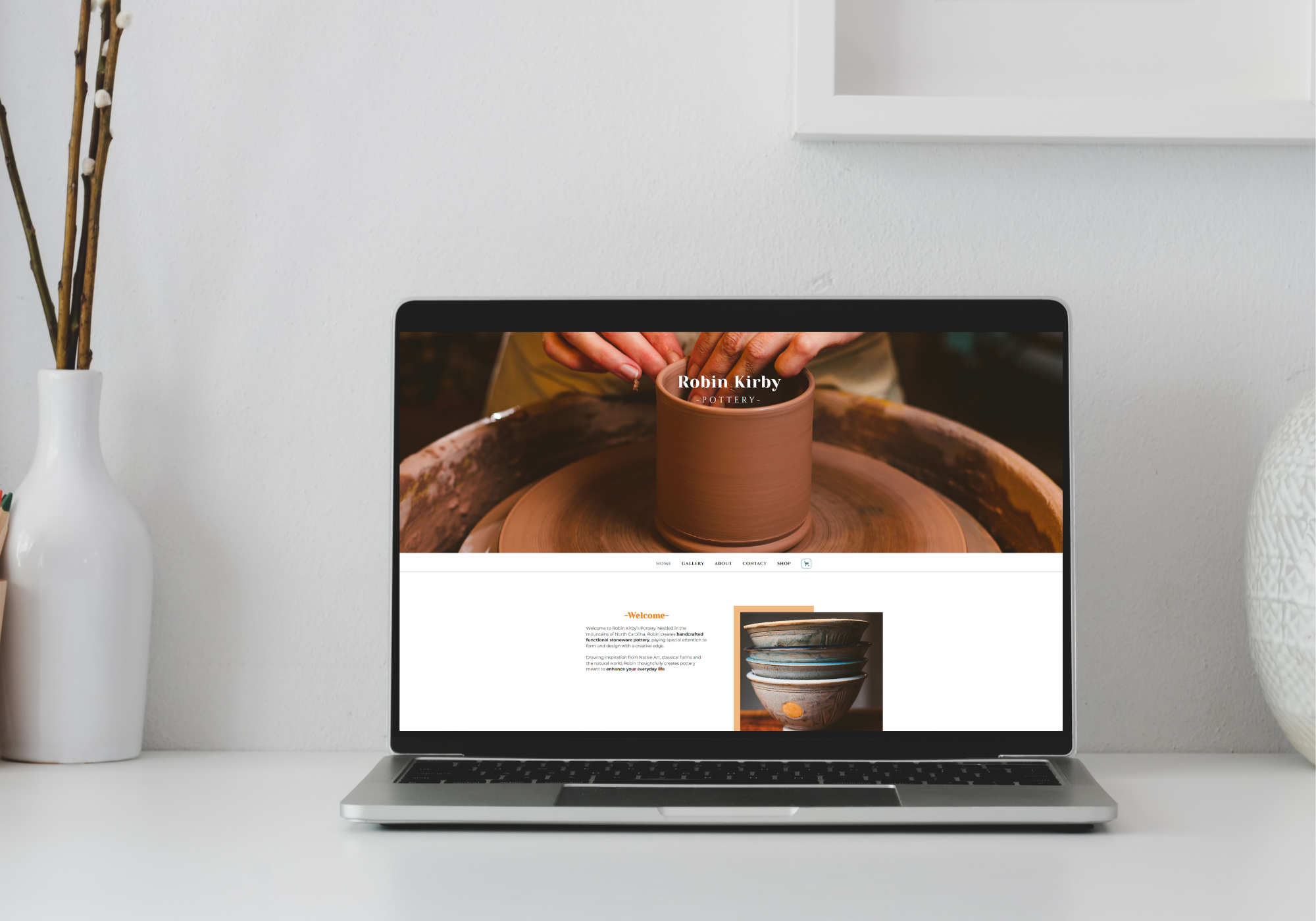 A picture of a desk with an open laptop, on the laptop is a screenshot of a potter's website, she is making a new pot on a potter's wheel. 