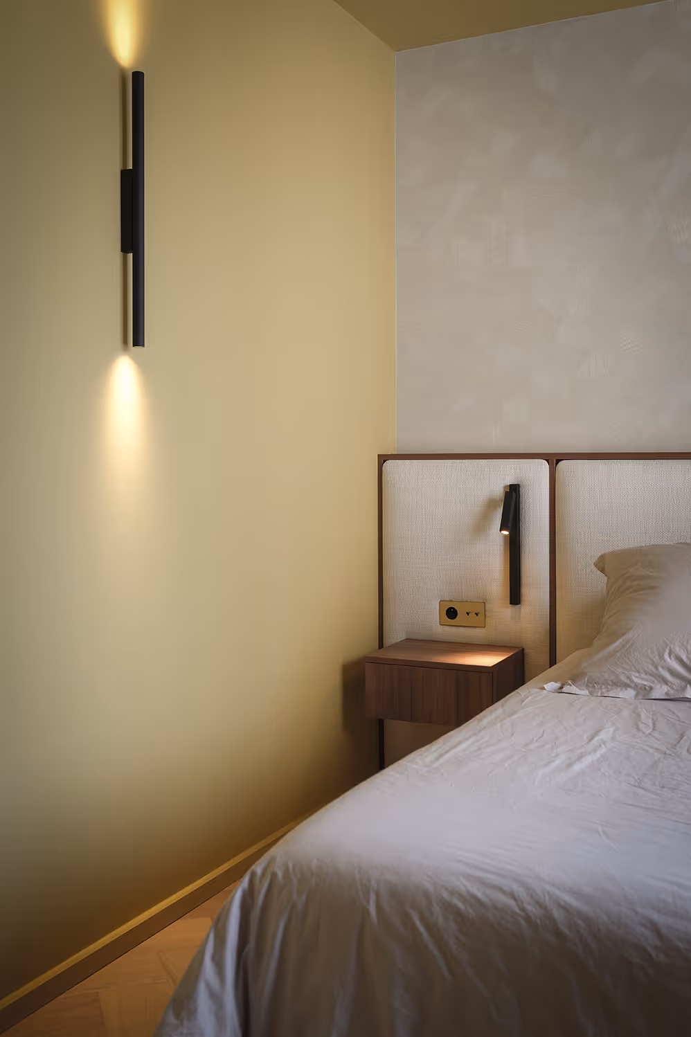 Chambre minimaliste avec mur jaune, tête de lit beige, table de chevet en bois et lampe murale noire allumée.