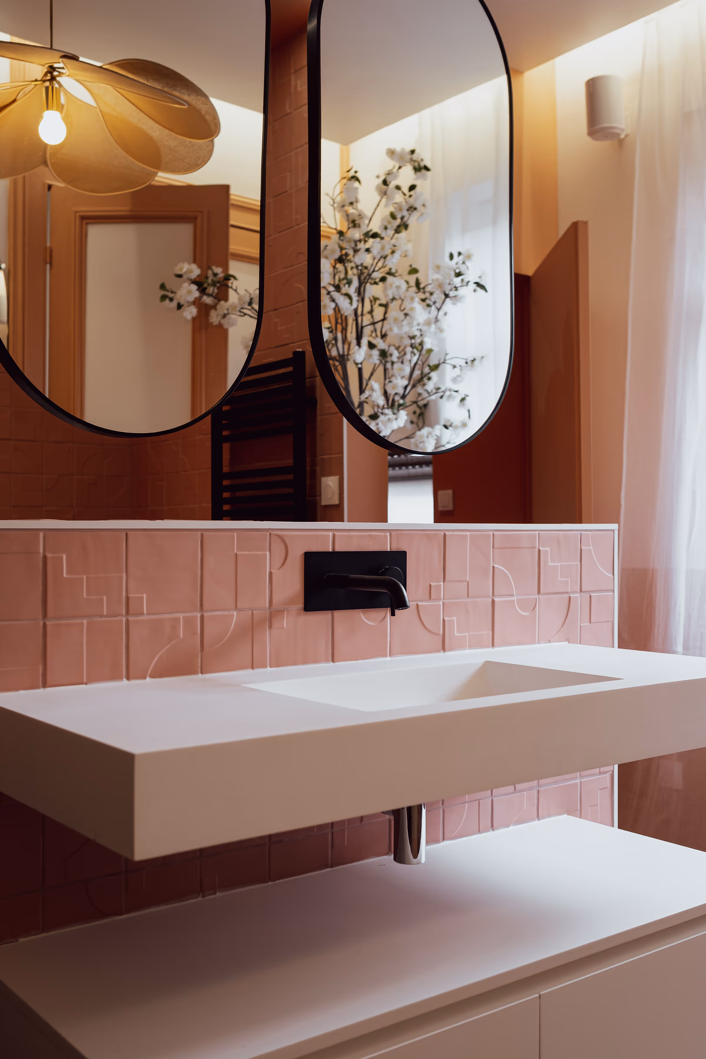 Lavabo blanc suspendu avec robinet noir et carreaux roses en arrière-plan, miroir ovale et lampadaire à fleur dorée.