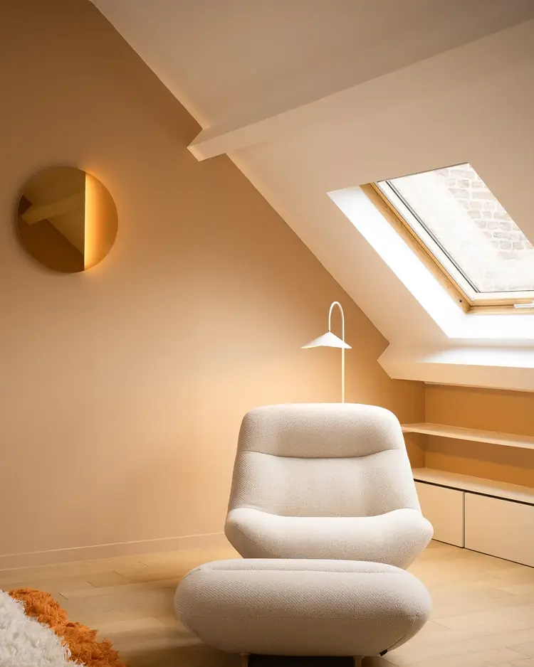 Coin salon sous combles d'un appartement à Lille : fauteuil et pouf en bouclé crème, murs terracotta, lampadaire arc et velux.
