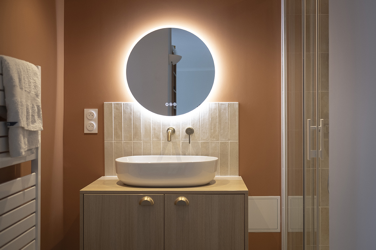 Salle de bain d'un appartement à Lambersart : vasque ovale sur meuble en bois clair, miroir rond rétroéclairé, robinetterie laiton et mur terracotta.