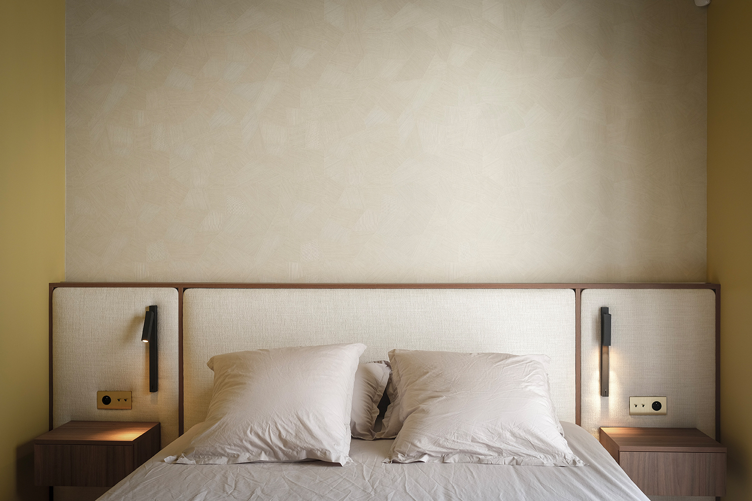 Chambre d'une maison à Téteghem : tête de lit capitonnée beige intégrée à une menuiserie en bois, liseuses noires et chevets assortis.