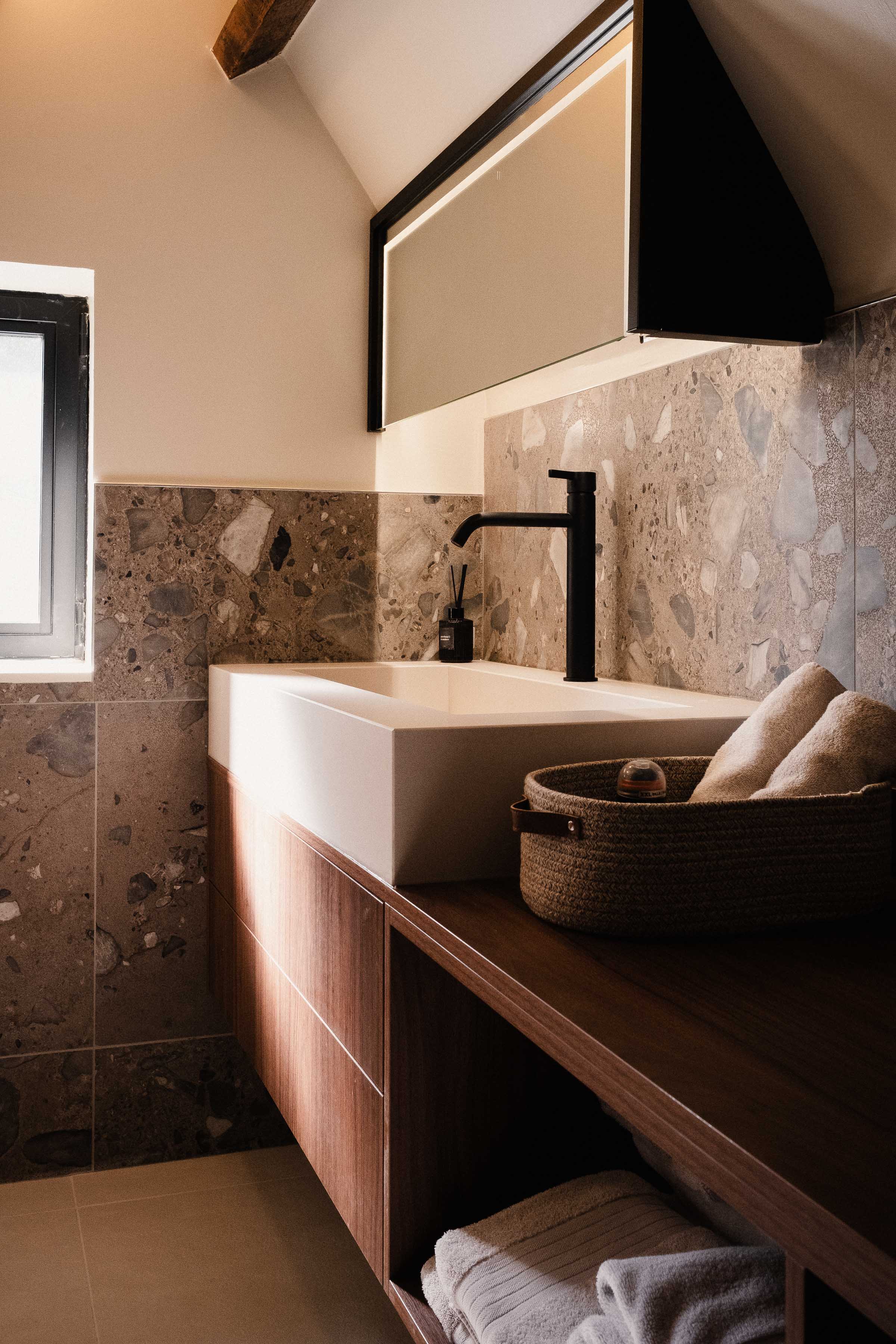 Salle de bain sous combles à La Madeleine : vasque blanche sur meuble bois, robinetterie noire, crédence en terrazzo et miroir rétroéclairé.
