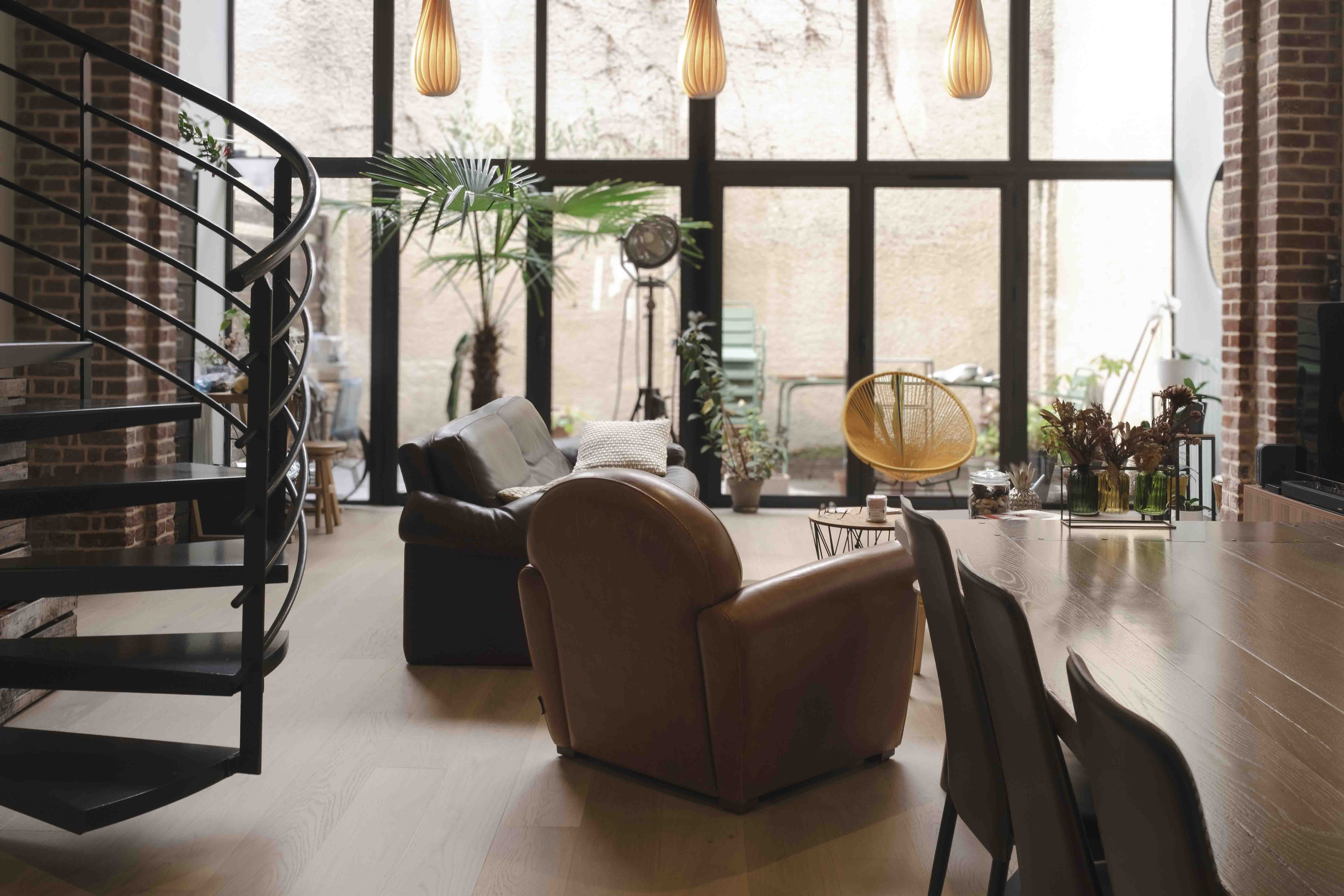Séjour d'un loft à Lille : verrière d'atelier en acier noir, escalier hélicoïdal, murs en brique et fauteuil club en cuir, sous double hauteur.