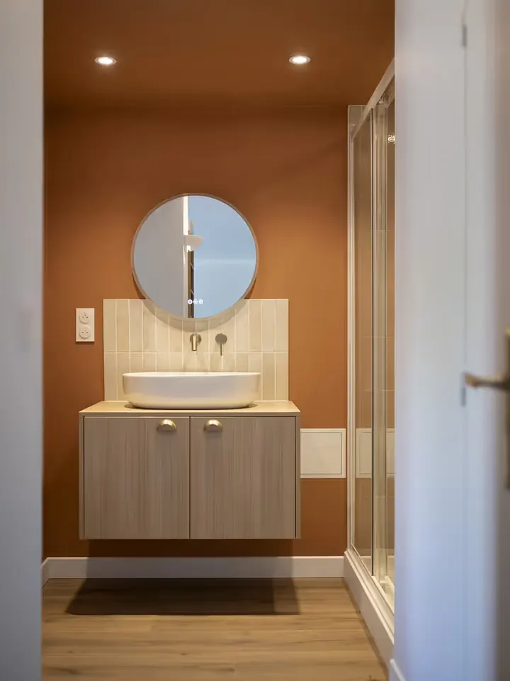 La salle d'eau livrée : meuble vasque en bois clair, miroir rond, mur terracotta et douche vitrée.