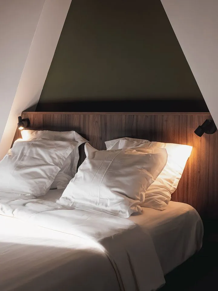 La chambre sous les combles livrée : tête de lit en bois, deux liseuses, mur vert sombre et linge de lit blanc.