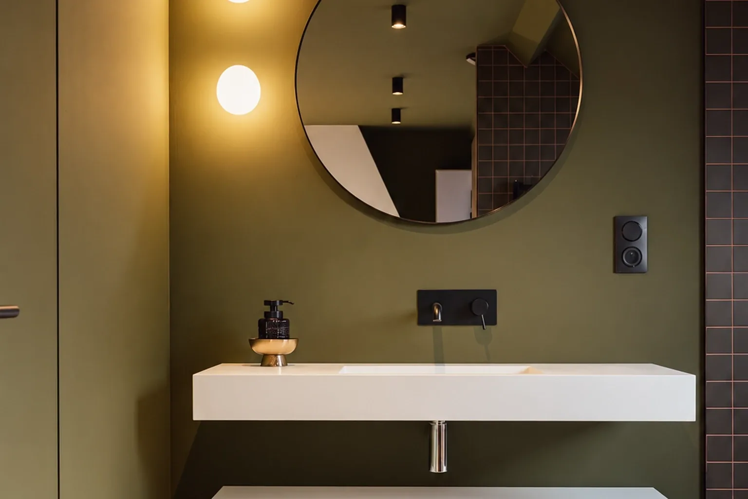 Vasque blanche suspendue et meuble blanc sous un miroir rond, avec deux appliques et un mur vert olive.