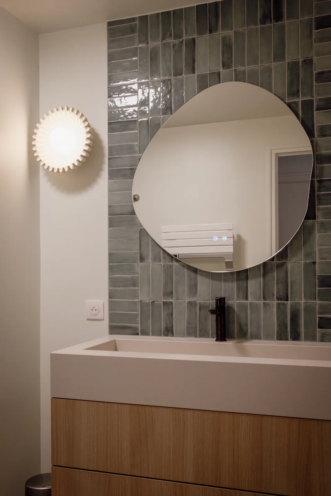 Meuble vasque en bois clair sous un miroir rond, devant un carrelage gris.