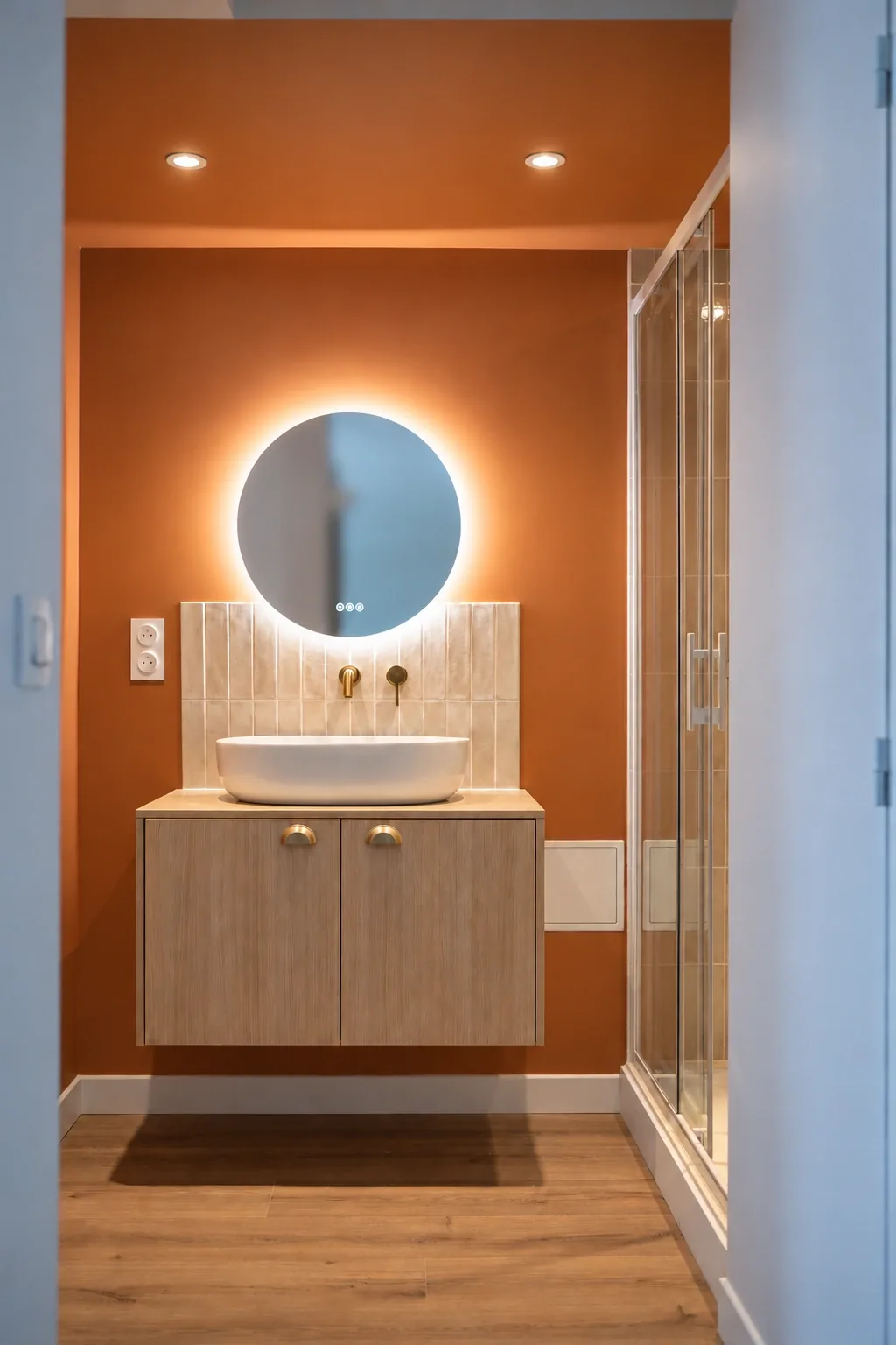 Perspective encadrée par une porte sur le meuble vasque, le miroir lumineux et la douche vitrée.