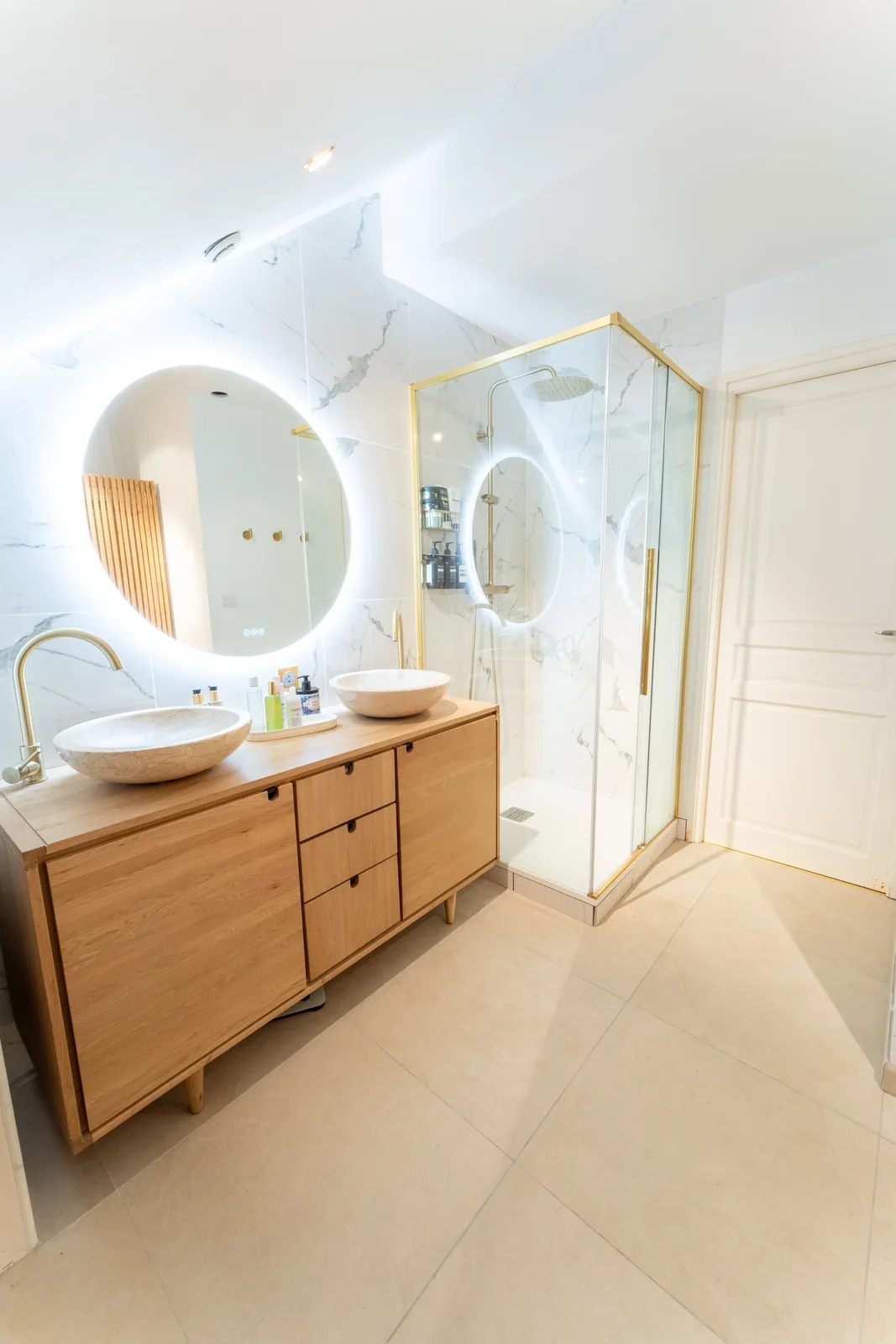 Meuble vasque en bois, miroir rond rétroéclairé et douche vitrée dorée dans une salle de bain blanche.