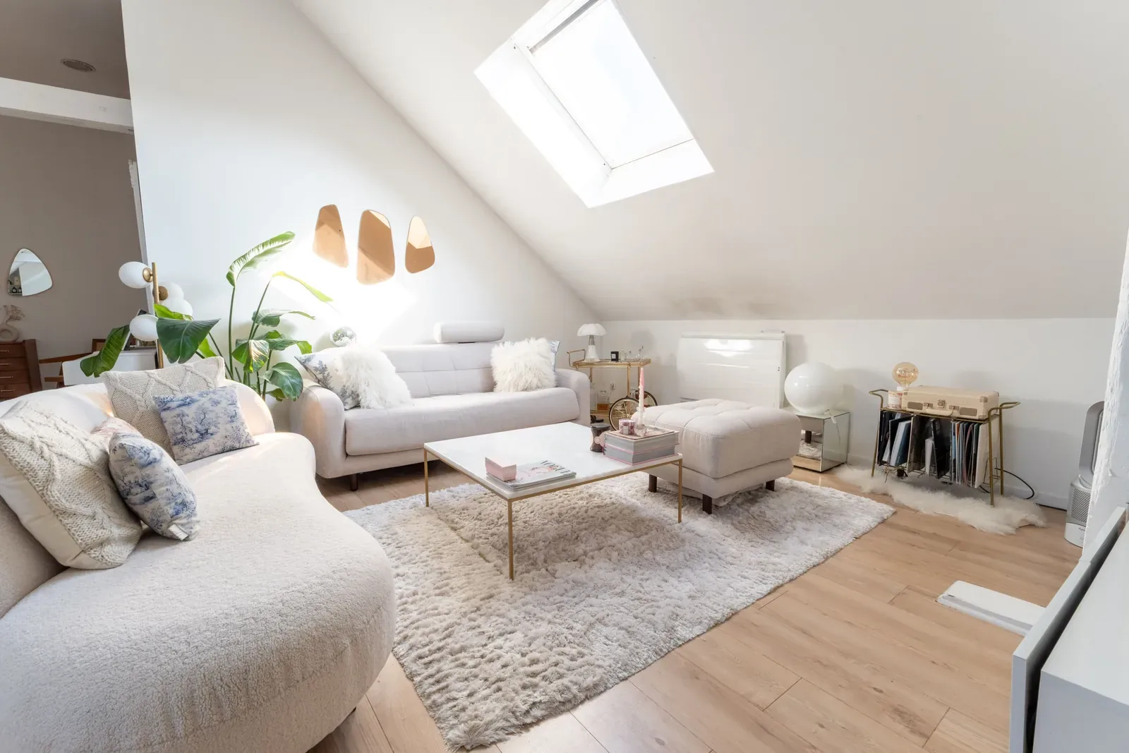 Salon lumineux d'un loft mansardé à Roubaix : canapé arrondi en bouclé blanc, table basse en marbre et laiton, grand tapis moelleux et velux.