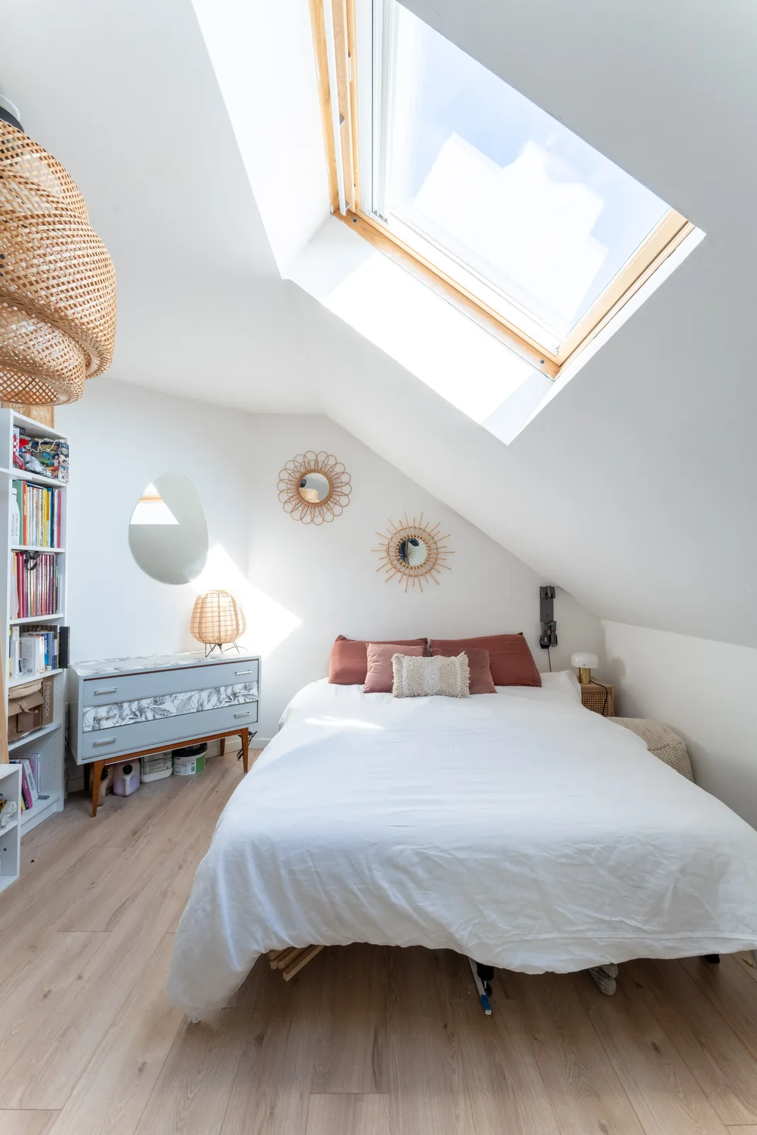 Chambre sous combles avec lit blanc, fenêtre de toit et suspension en rotin.