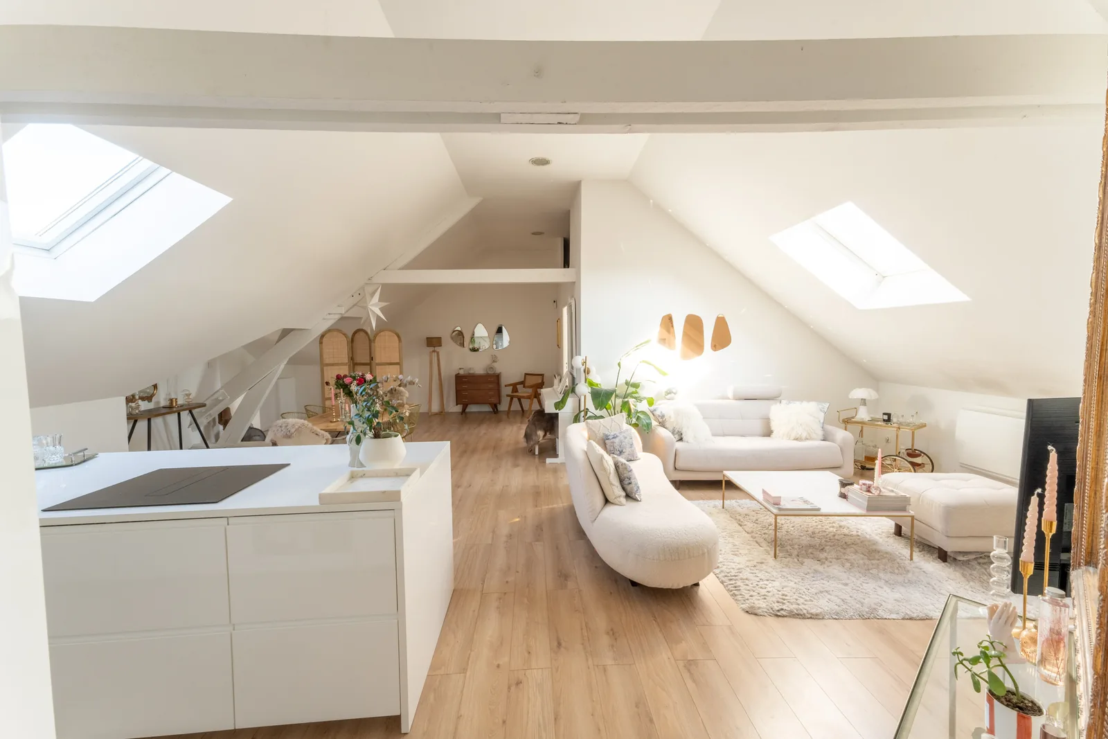 Loft sous combles : îlot de cuisine blanc ouvert sur le salon, poutres peintes en blanc et fenêtres de toit.