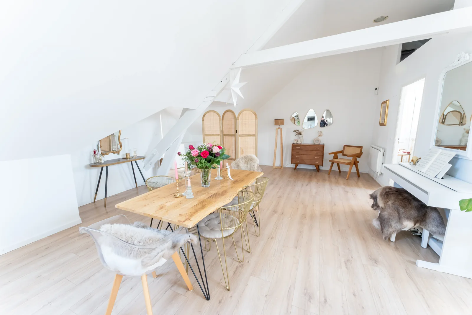Salle à manger du loft, table en bois clair et chaises en rotin sous les poutres blanches.