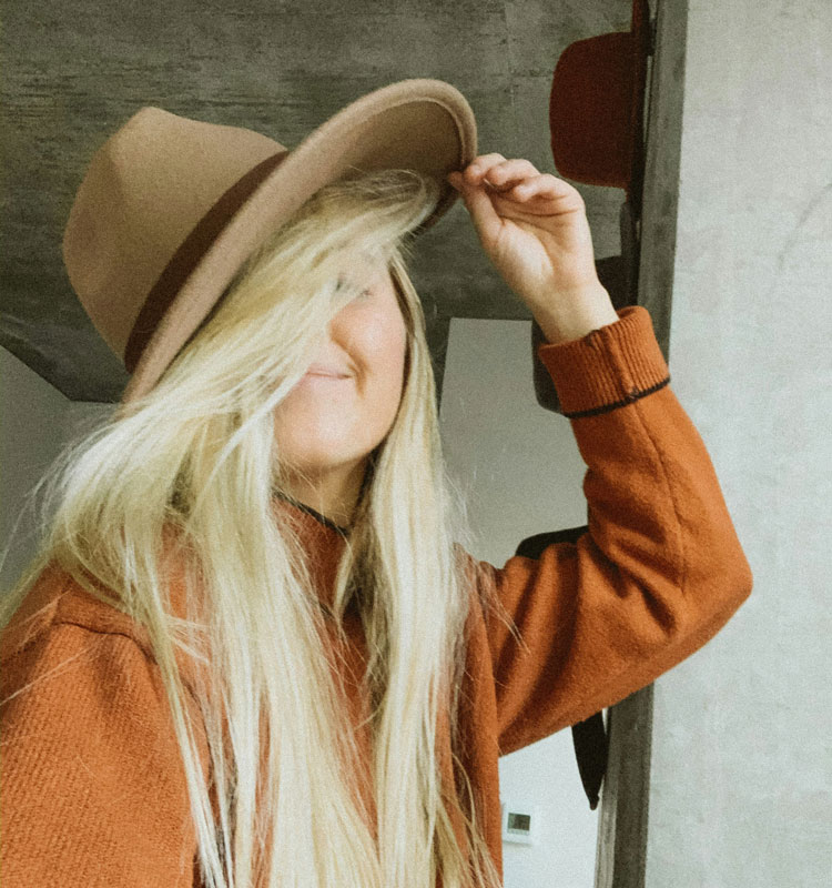 Femme blonde souriante portant un chapeau en feutre beige, vêtue d’un pull orangé, dans un intérieur chaleureux et minimaliste.