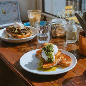 Deux assiettes de brunch avec œufs pochés, toasts et pesto sur une table en bois près d’une fenêtre ensoleillée chez MILO.