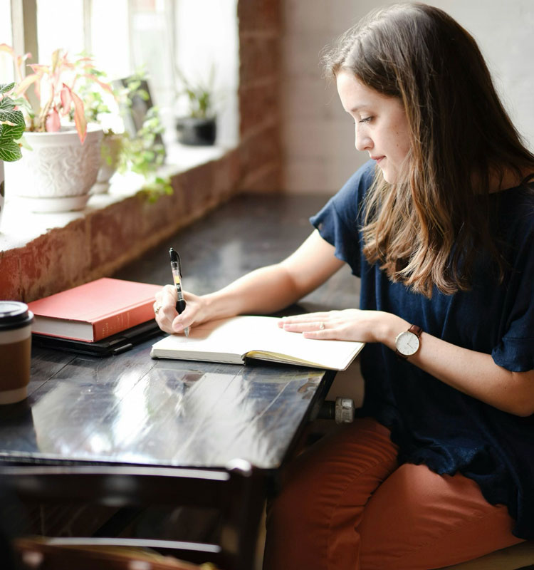 Femme concentrée en train d’écrire dans un carnet sur une table près d’une fenêtre, dans une ambiance lumineuse et studieuse.