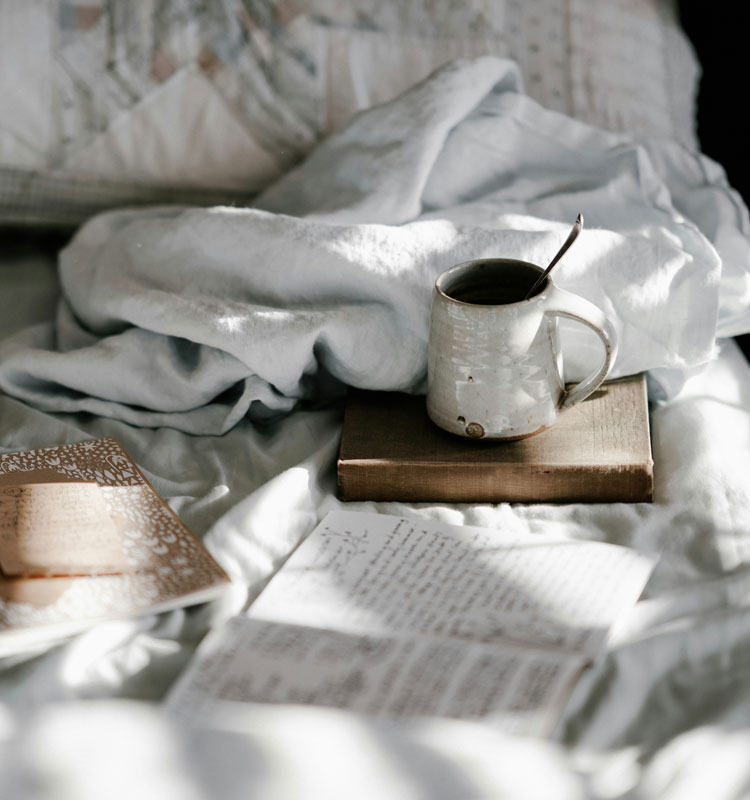 Tasse en céramique posée sur un livre, sur un lit recouvert de draps blancs froissés, accompagnée de carnets et de feuilles manuscrites, baignée par une lumière douce du matin.