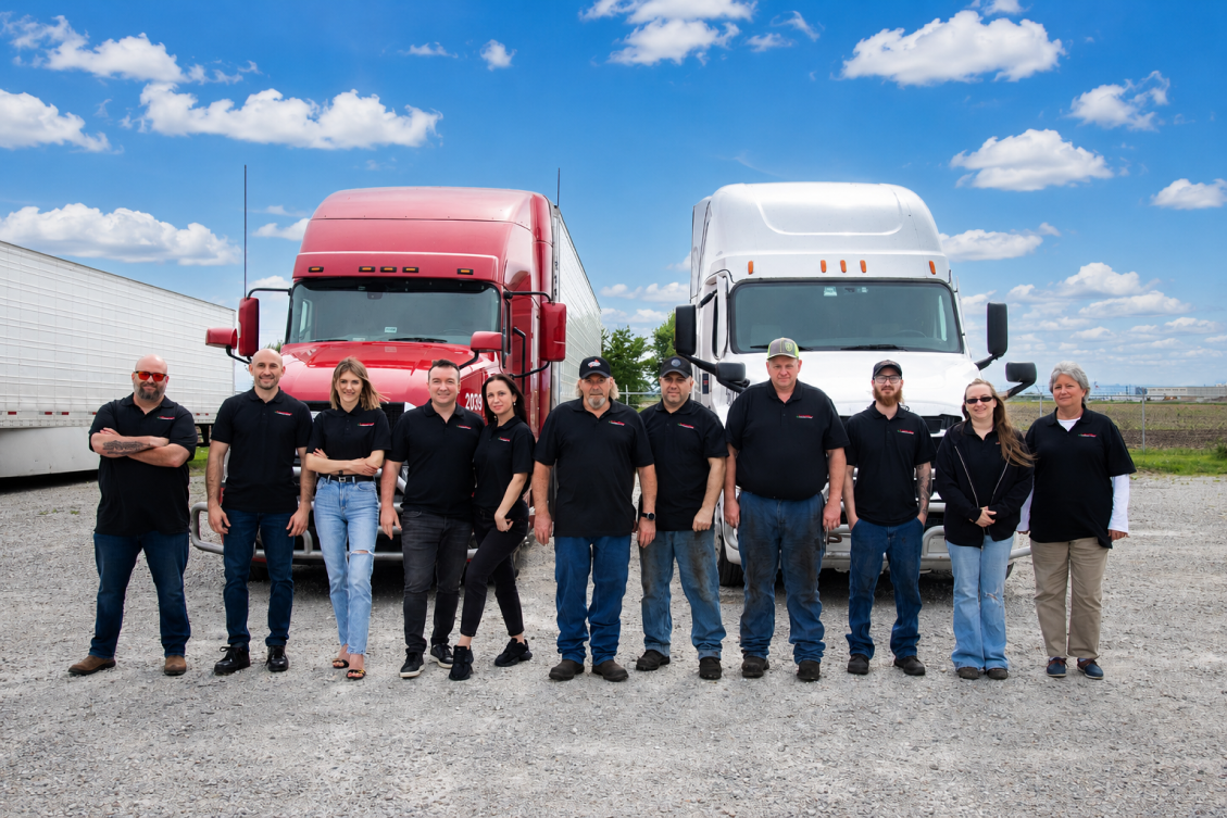 Group of ten people standing in front of red and white semi trucks on a gravel lot under a blue sky.