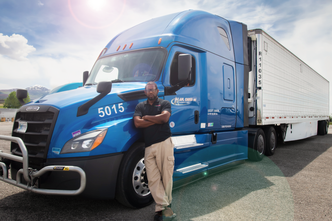 Man with arms crossed leaning against blue semi-truck tractor in a sunny parking lot.