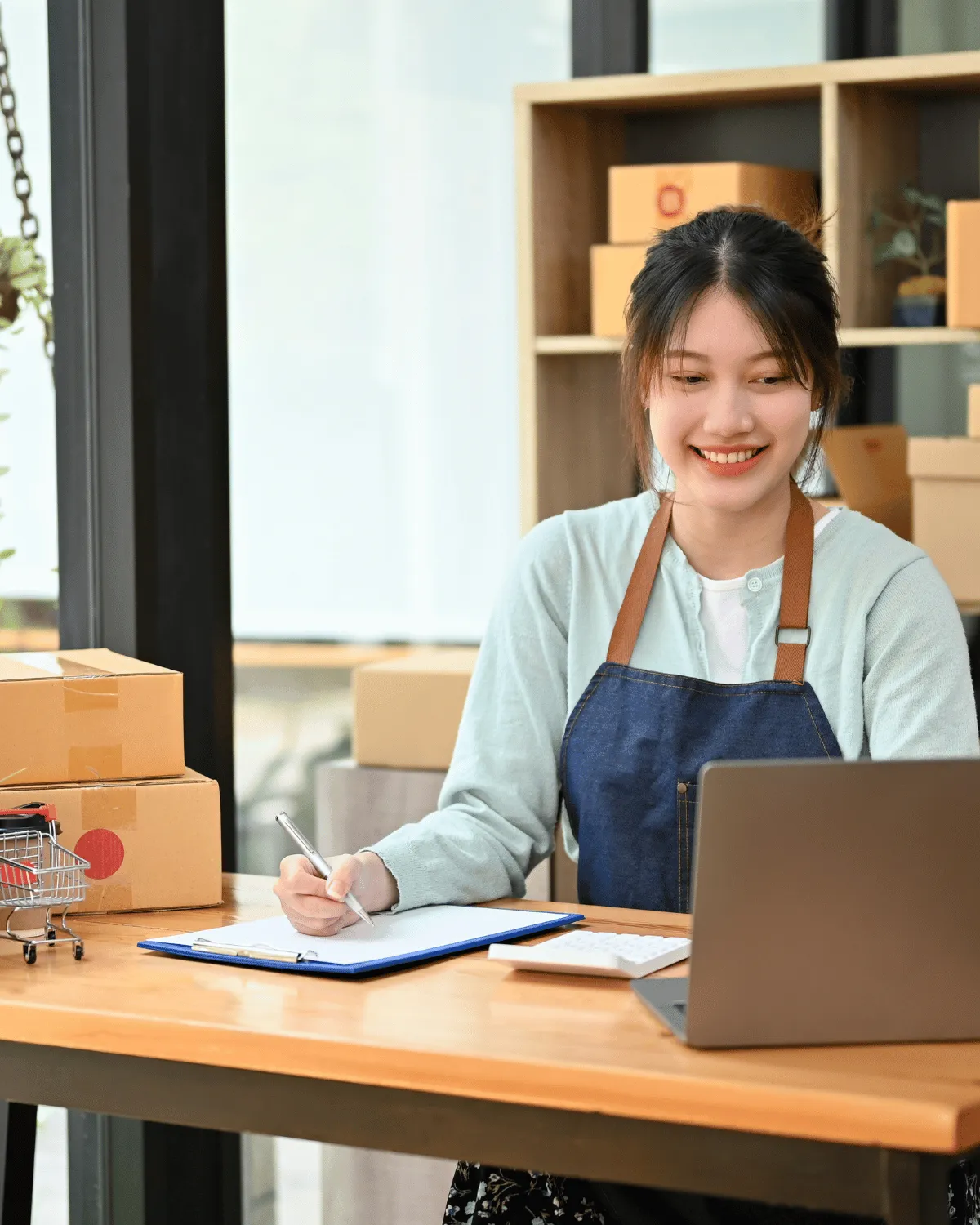 Jeune femme souriante en tablier utilisant un ordinateur portable et prenant des notes à un bureau entouré de cartons d'expédition.