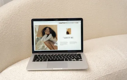 Ordinateur portable ouvert affichant une page web 'About Me' avec la photo d'une femme portant un chapeau de paille, posé sur un canapé beige texturé.