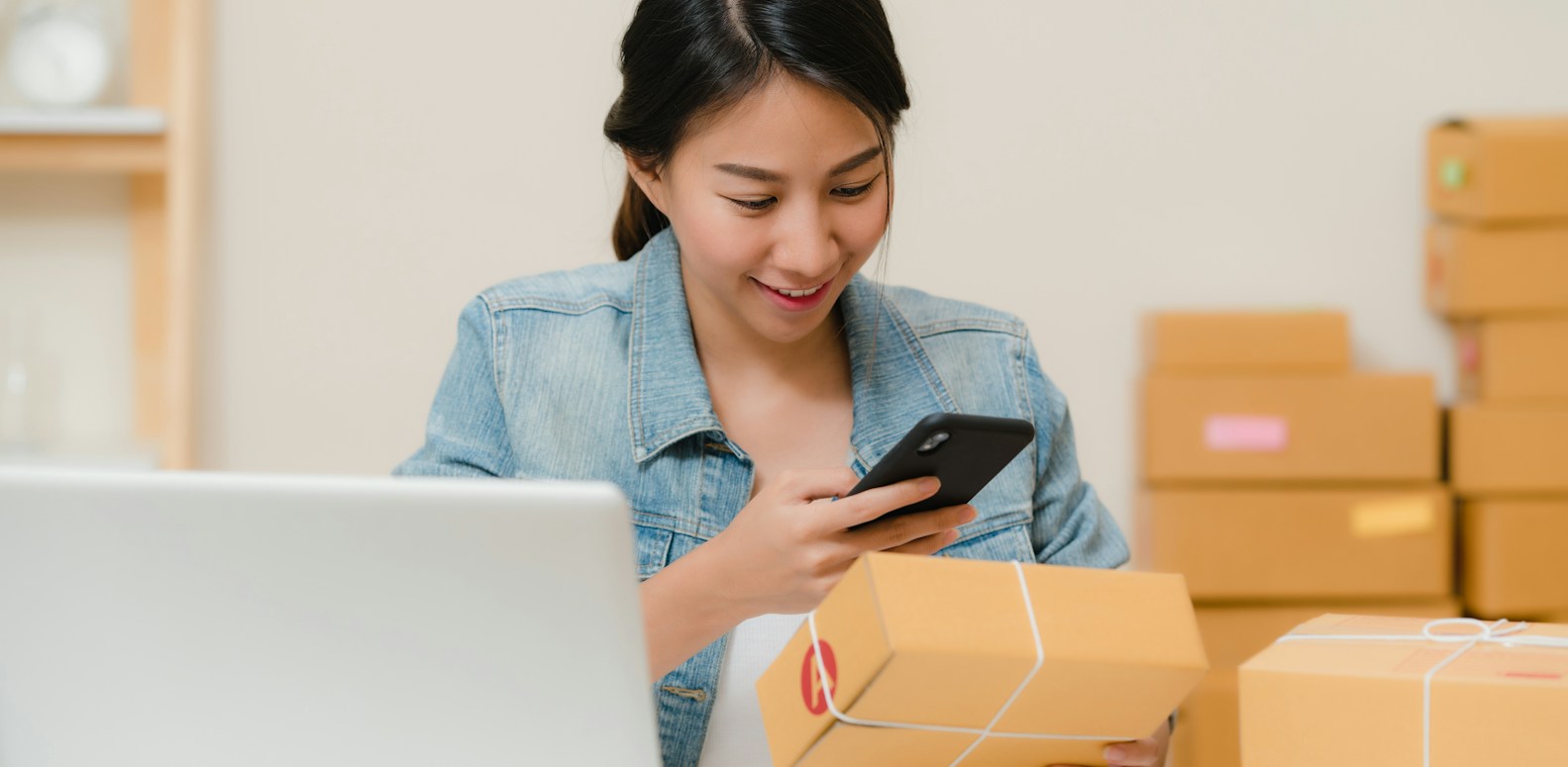Entrepreneuse vérifiant un colis avec son smartphone à côté d'un ordinateur portable et de cartons