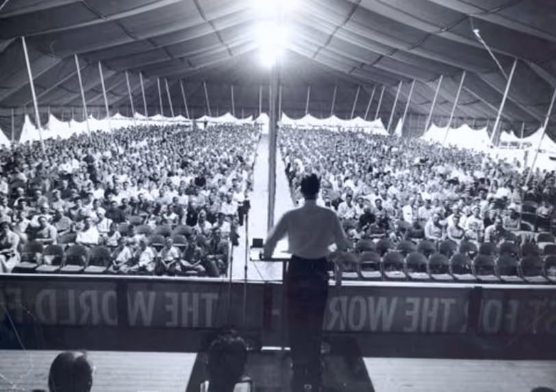 Large tent revival meeting with a preacher addressing a packed audience