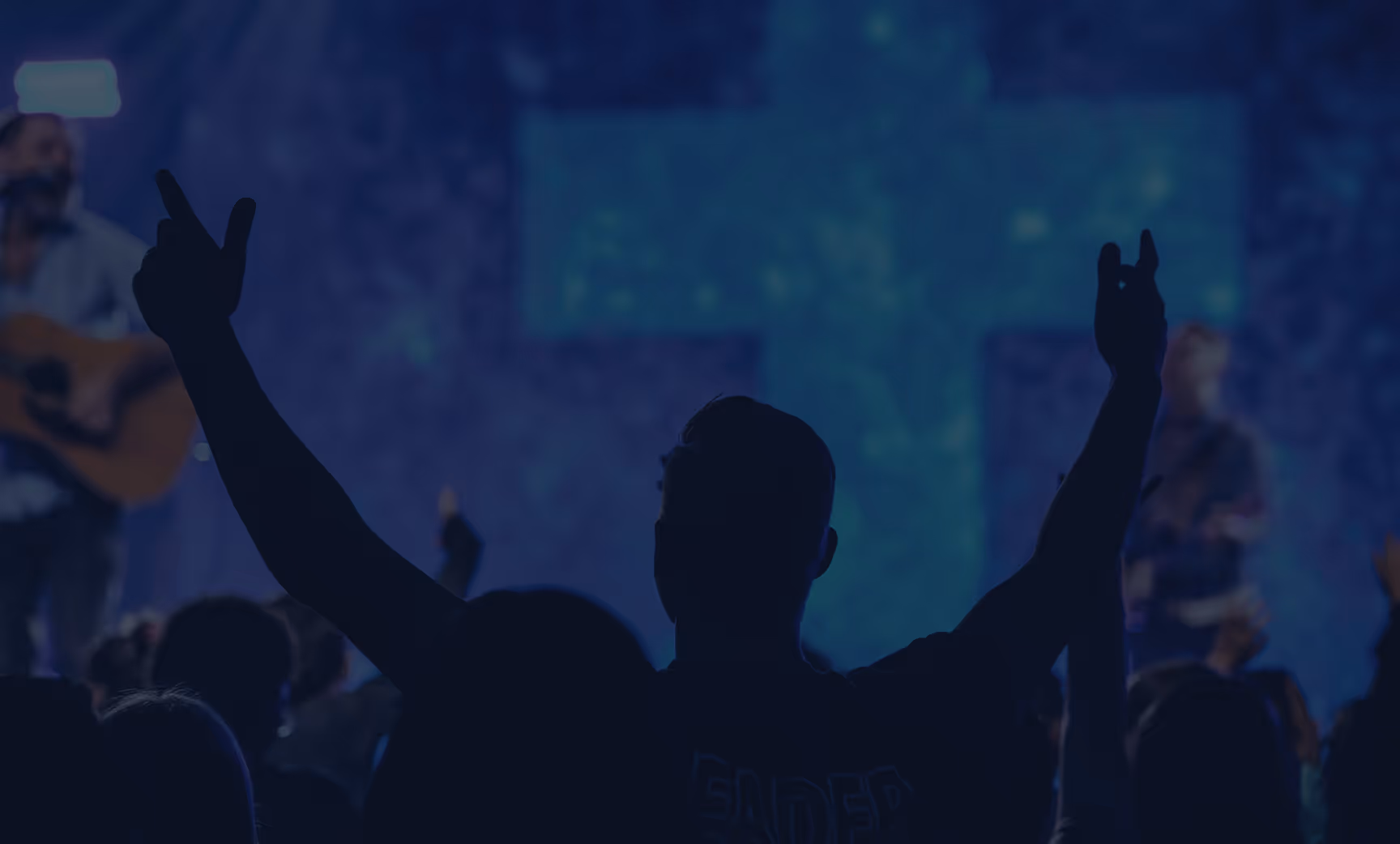 Silhouette of a person worshipping with raised hands during a live Christian music performance, with a large illuminated cross in the background.