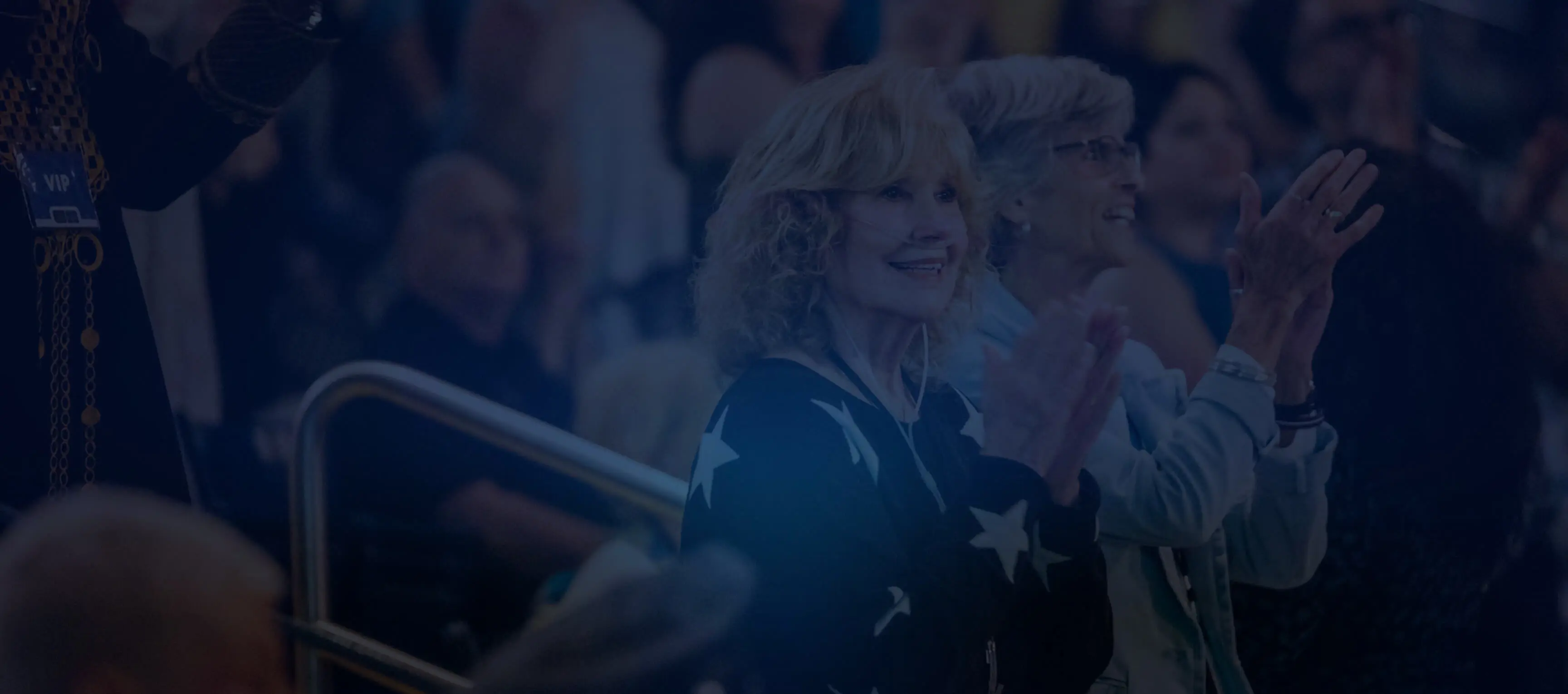 Two older women smiling and clapping at an event with a crowd in the background.