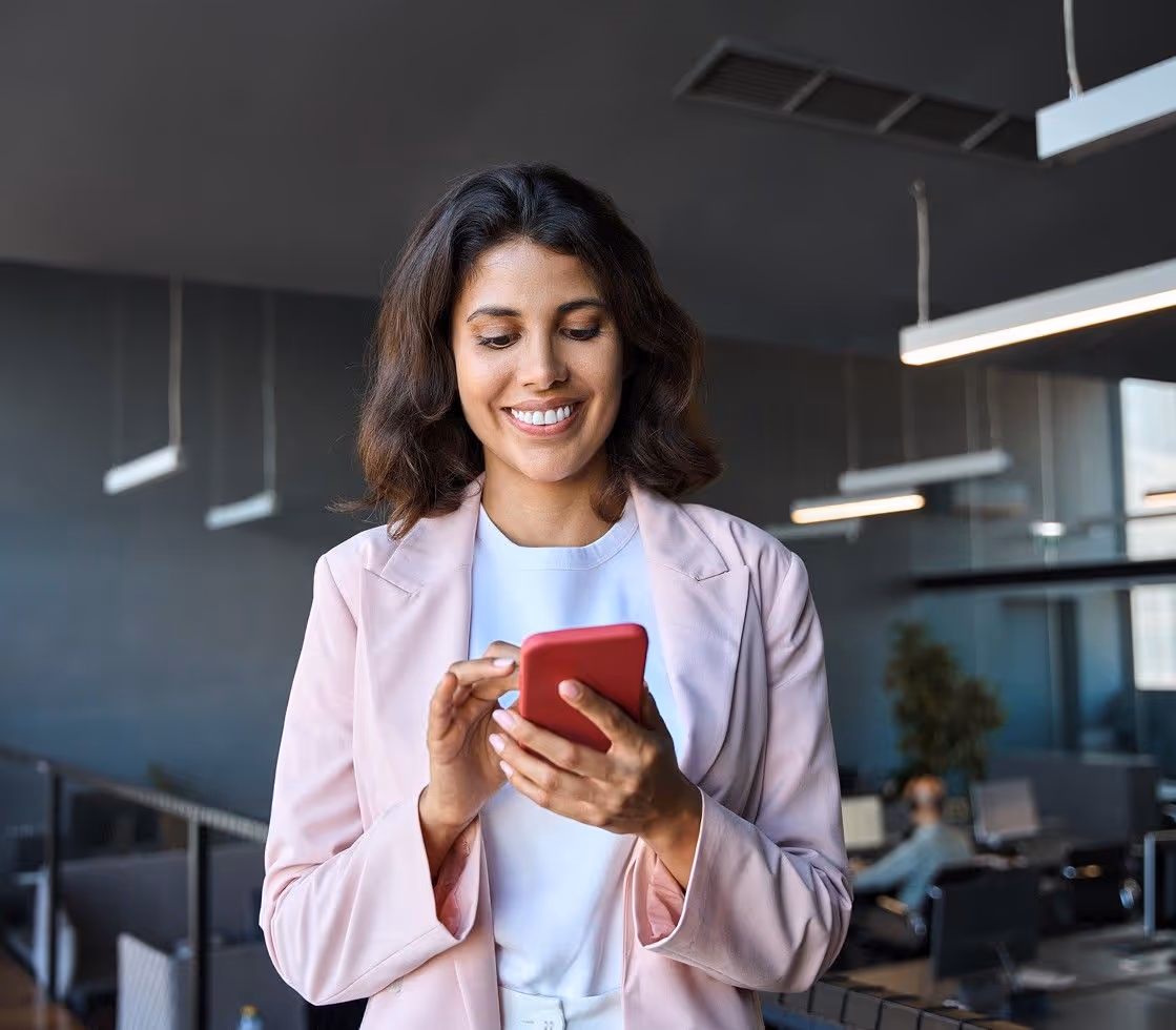 Smiling woman in white shirt and pink blazer using a red smartphone in a modern office.