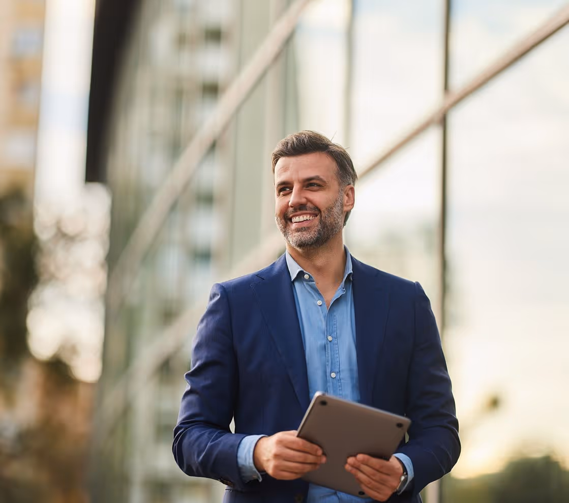 Smiling businessman in navy blazer holding tablet outdoors near glass building.