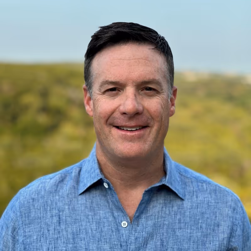 Smiling man with short dark hair wearing a blue button-up shirt outdoors with green blurred background.