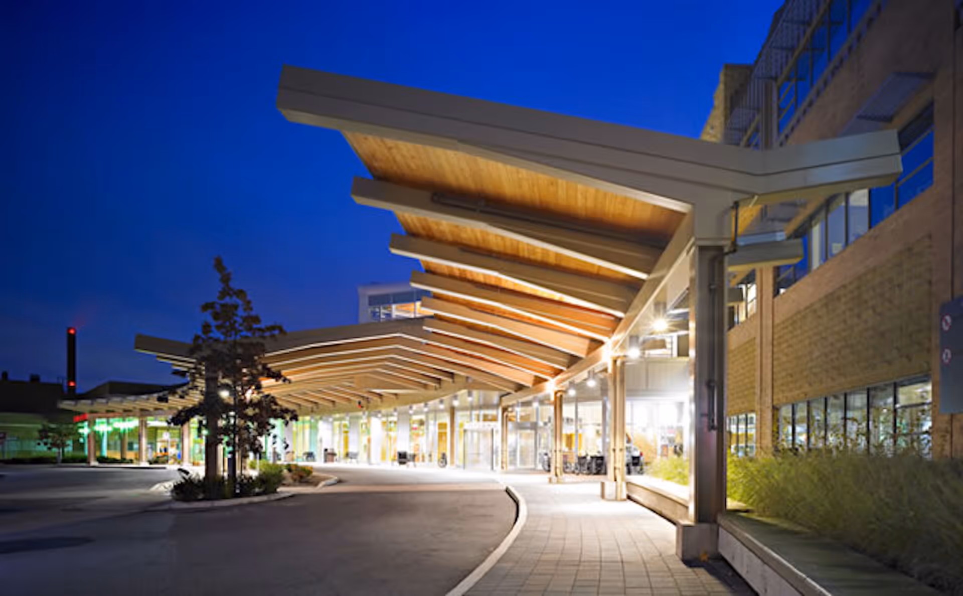 Building exterior at dusk with illuminated wood canopy and glass entrance along a curved driveway.