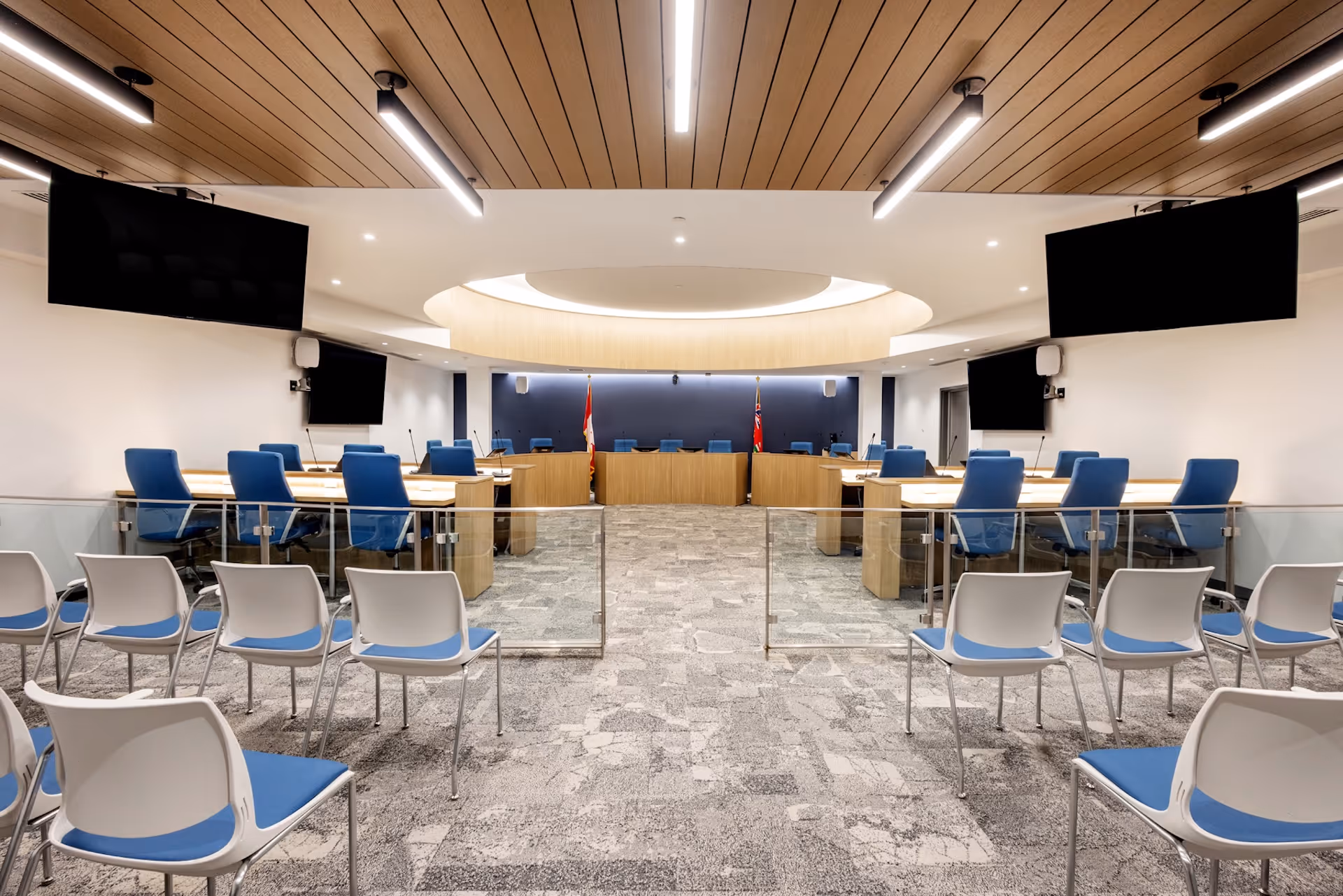 Modern council chamber interior with blue seating, wood ceiling panels, and a circular light feature above the meeting desks.