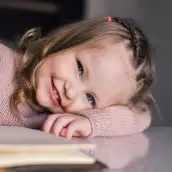 Момиче с плитки и розов пуловер, легнало върху маса и усмихващо се.