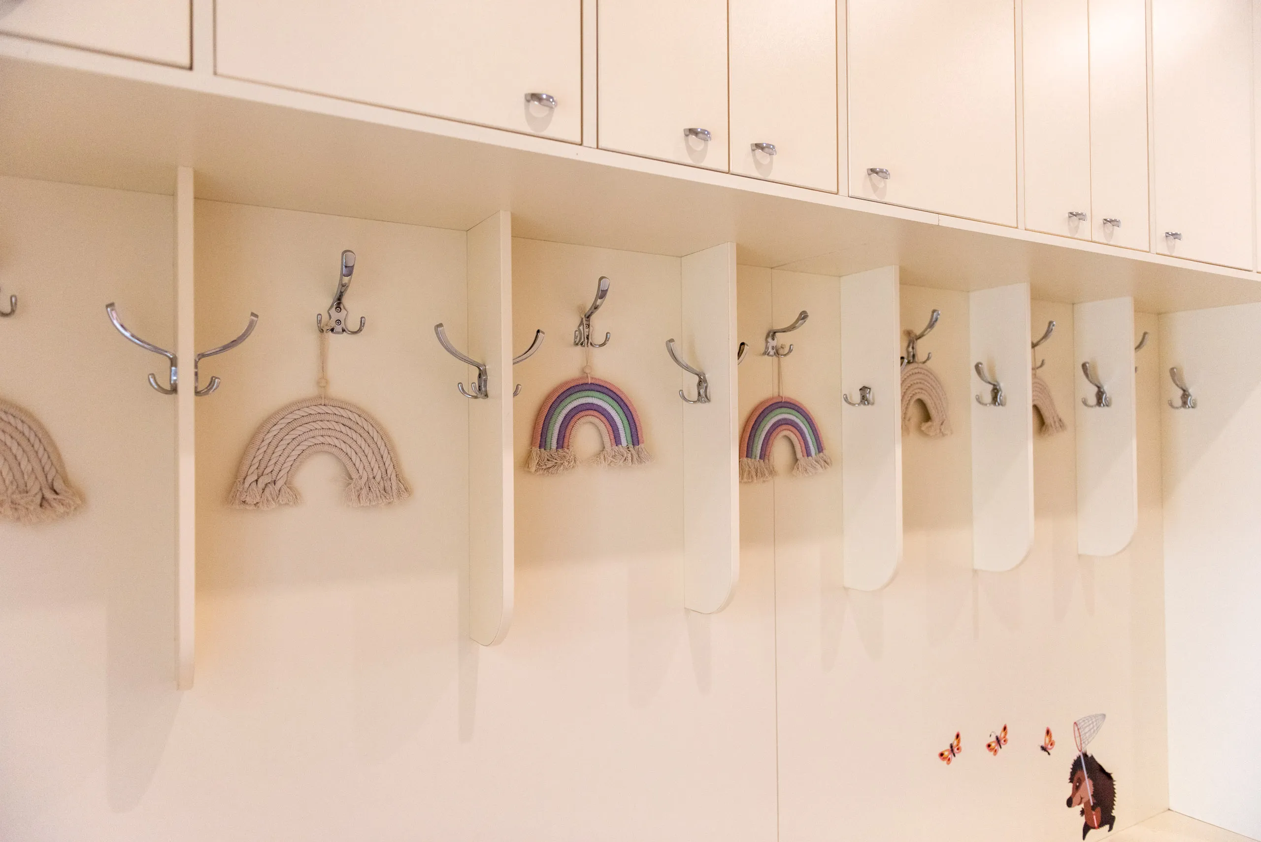 Бели шкафчета с метални закачалки, от които висят плетени дъговидни декорации и стикери на пеперуди и таралеж с мрежа за ловене.