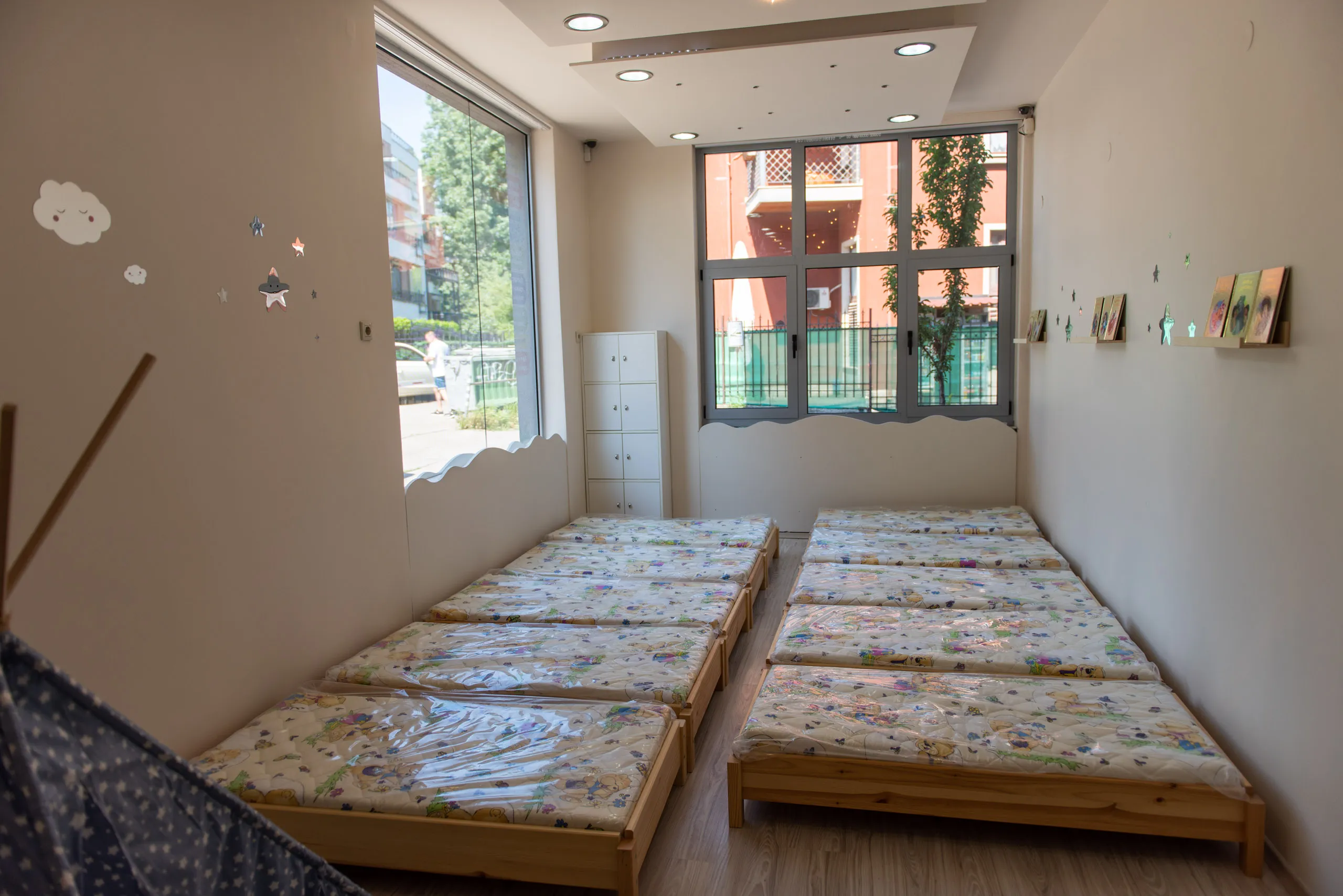 Детска спалня с осем малки легла, разположени в два реда, с прозорци и декорации на стени.