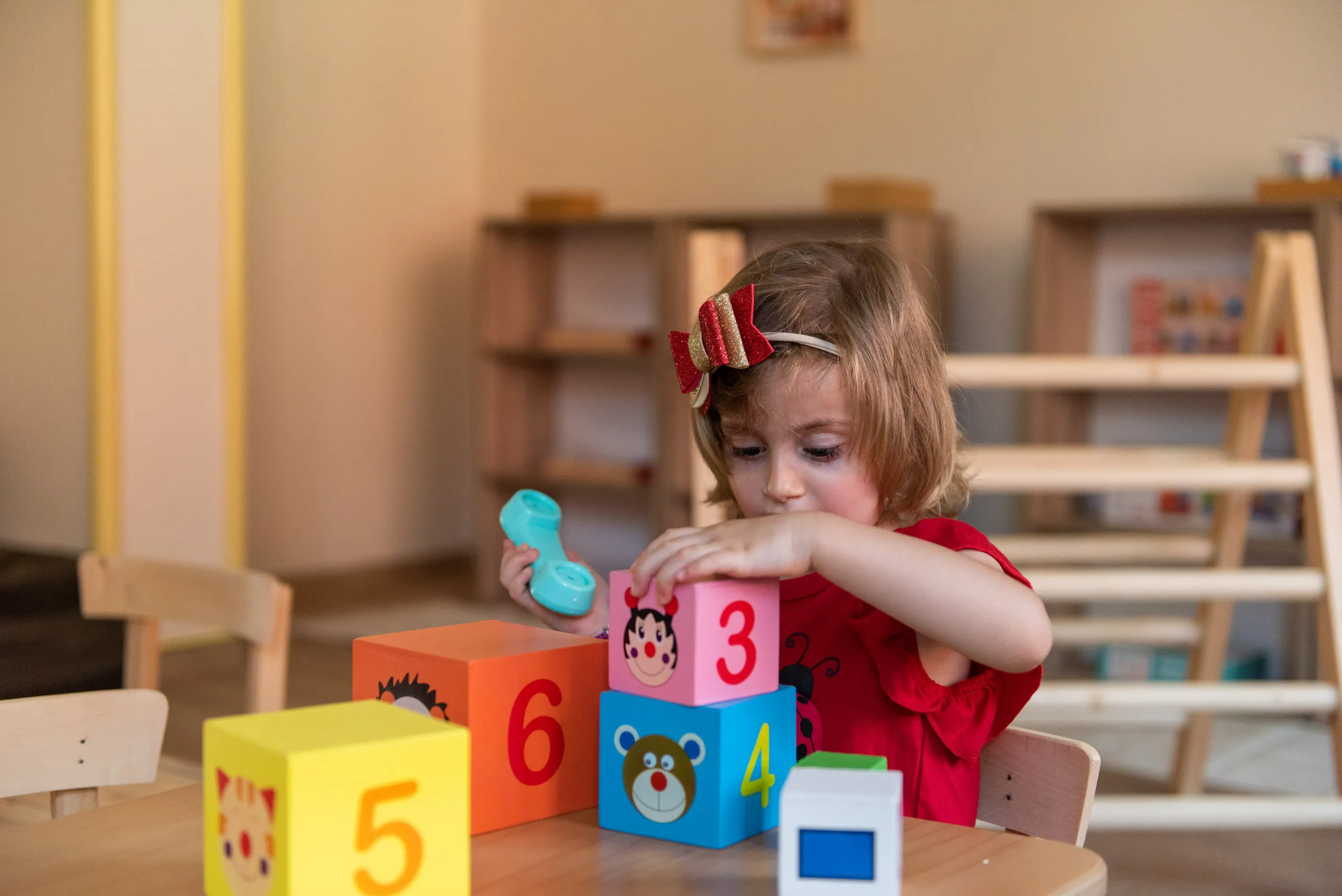 Малко момиче с червена лента играе с цветни строителни кубчета с номера и държи синя играчка телефон.