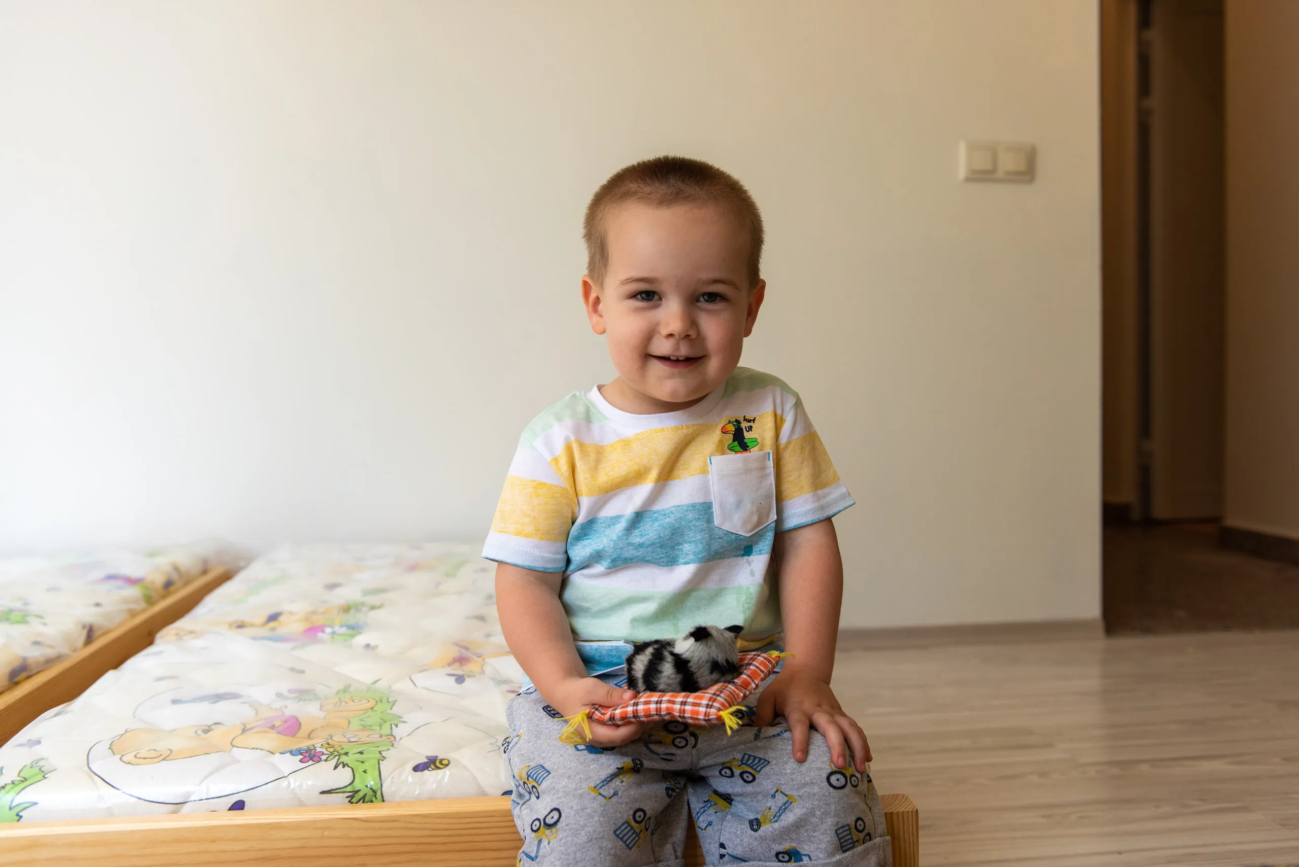 Малко момче, седящо на легло в стая, държи малък черно-бял пухкав домашен любимец върху оранжеви квадратни възглавнички.