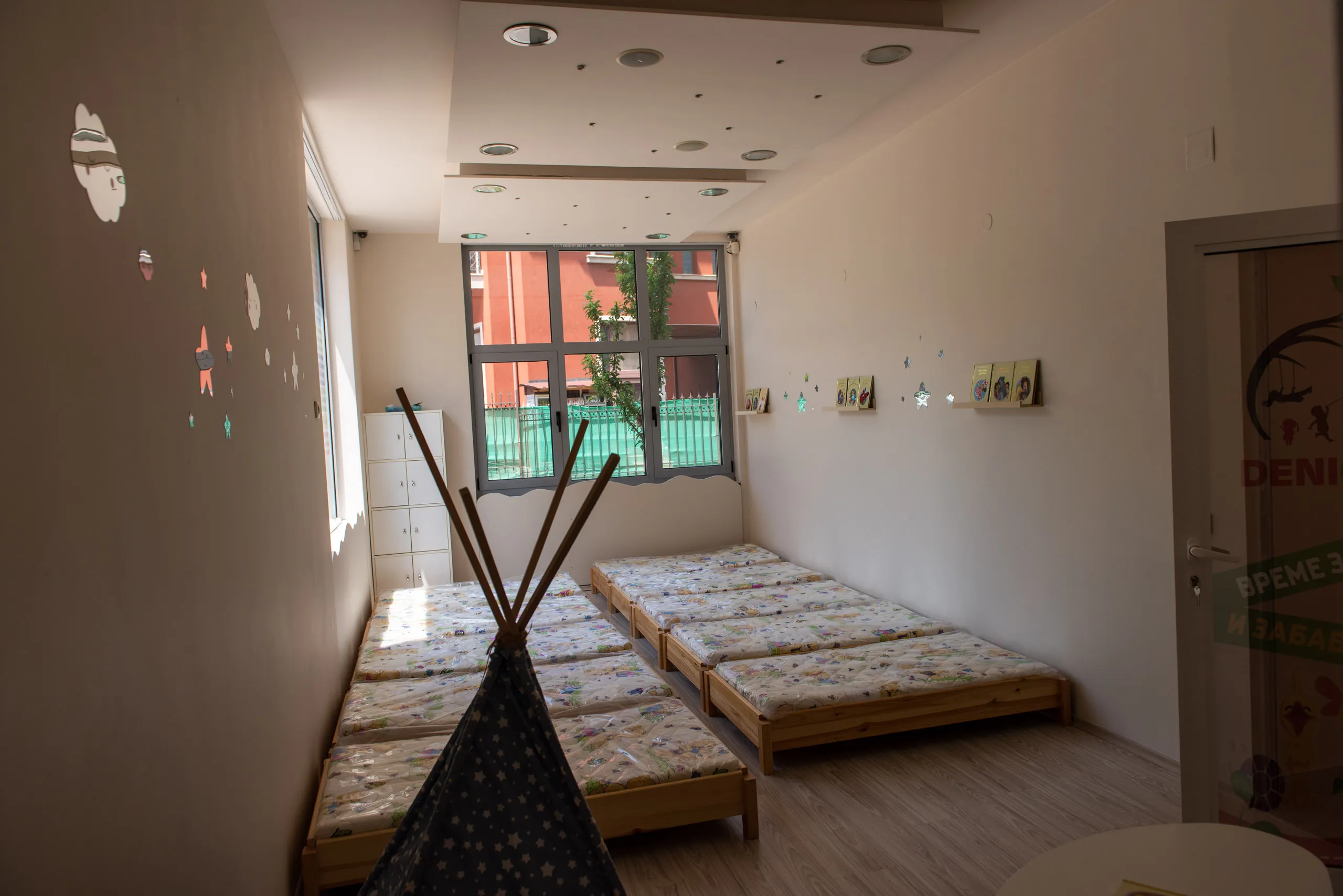 Детска стая с няколко единични легла с матраци с шарки, малка библиотека на стената и детски шатър в преден план.