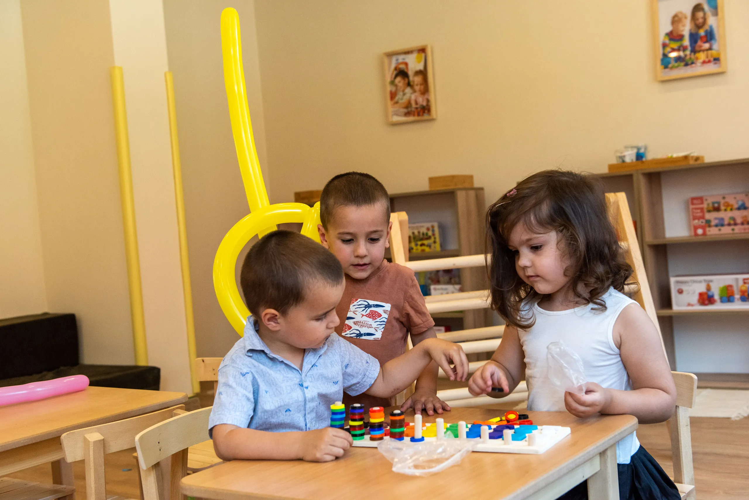 Три деца бележат игра докато седят на малки маси в класна стая с детски играчки и балони.