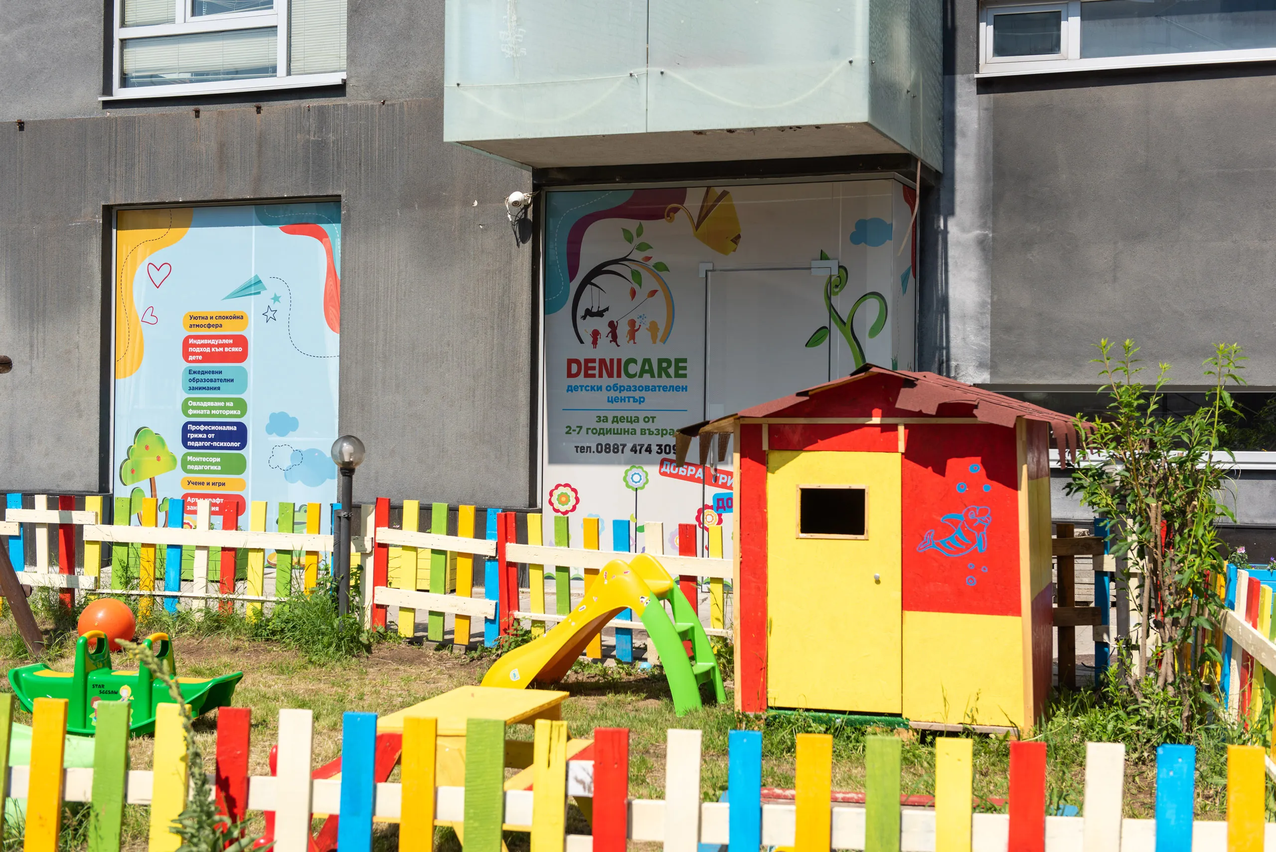 Ярка детска площадка с малка пързалка и цветна ограда пред детски образователен център.