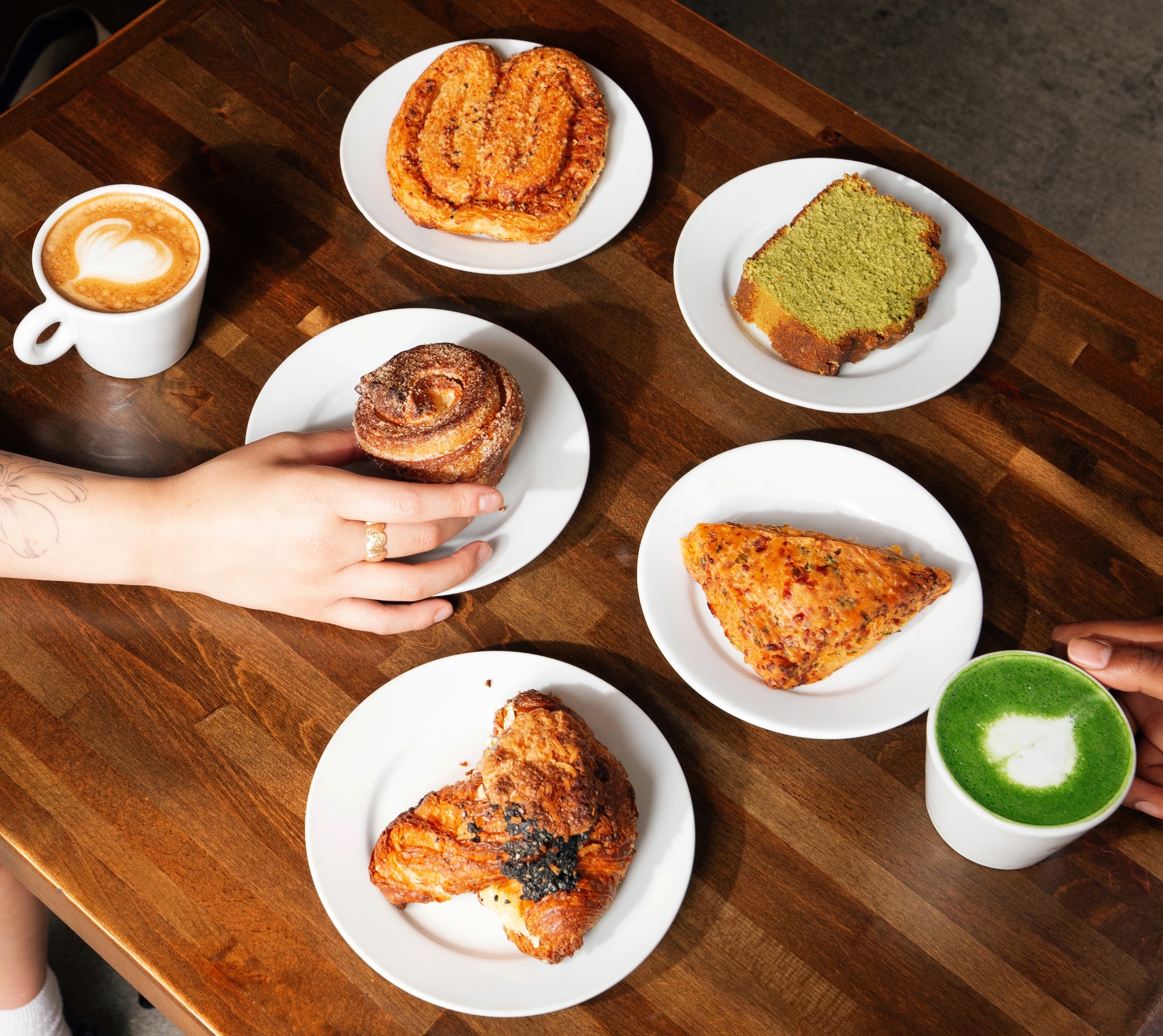 Two hands reach for cinnamon roll and matcha latte among assorted pastries and coffee on a wooden table.