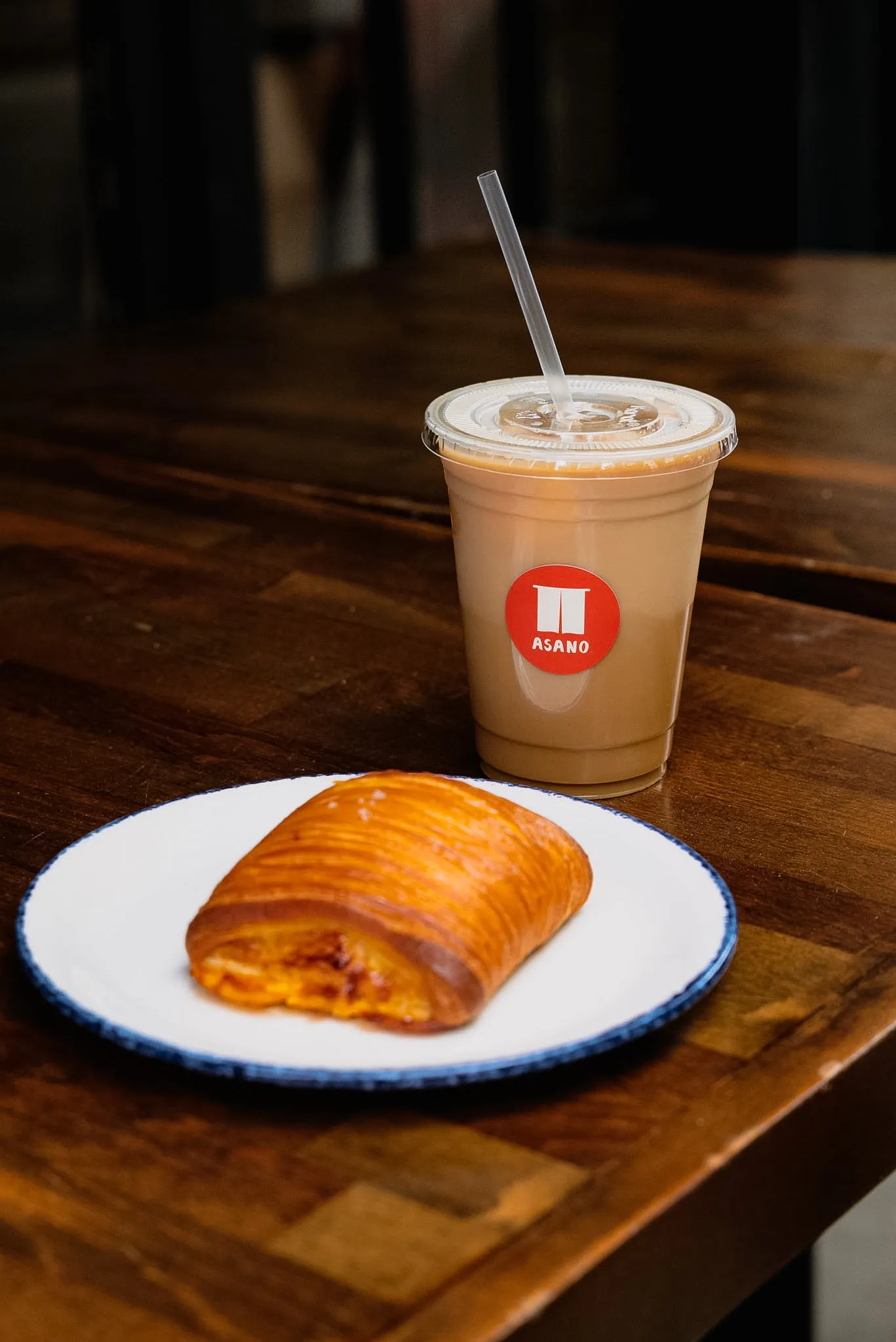 A laminated pastry on a blue-rimmed plate next to an iced latte with the ASANO logo