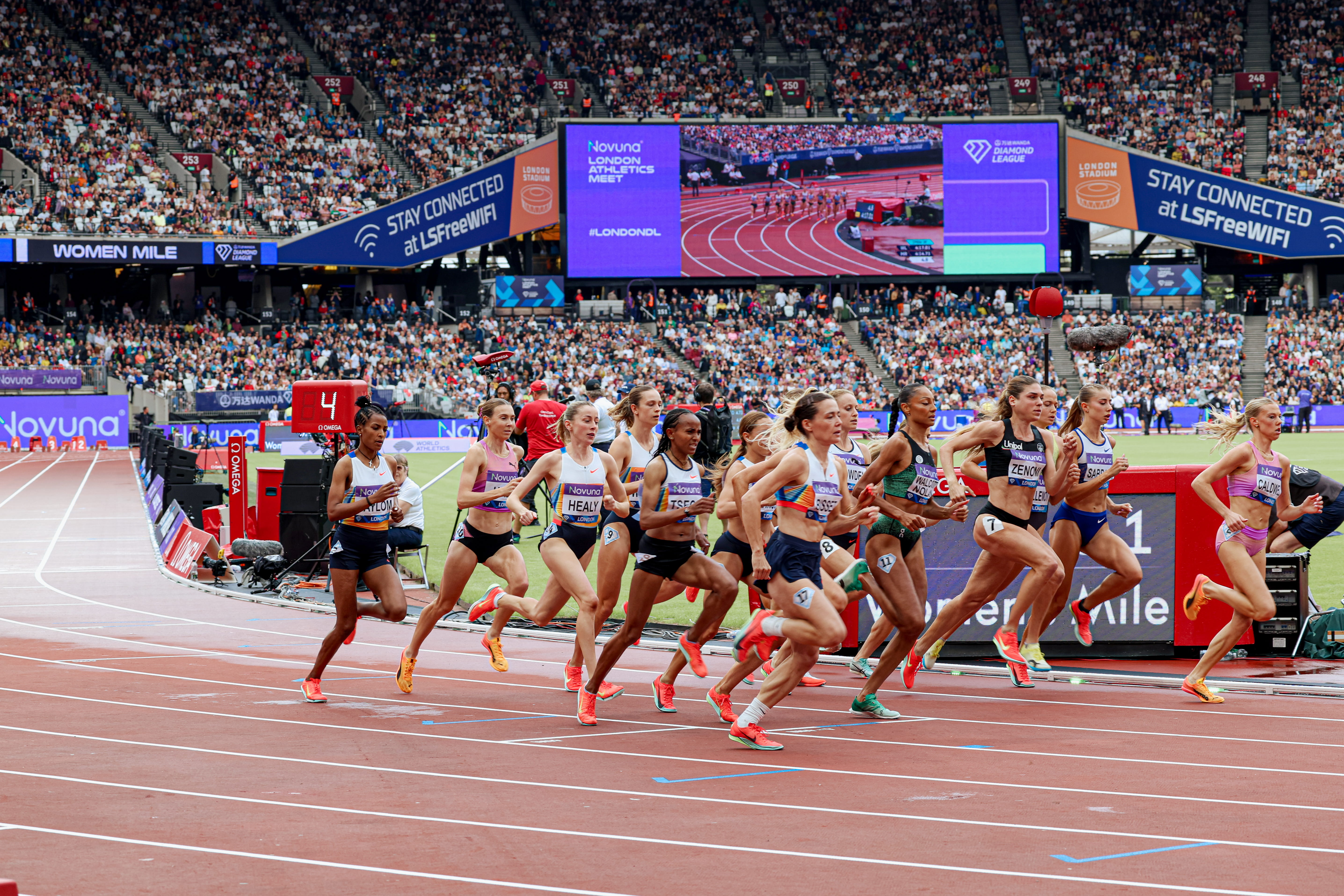 Female athletes sprinting on track during the women’s mile race at London Stadium with a large crowd watching.